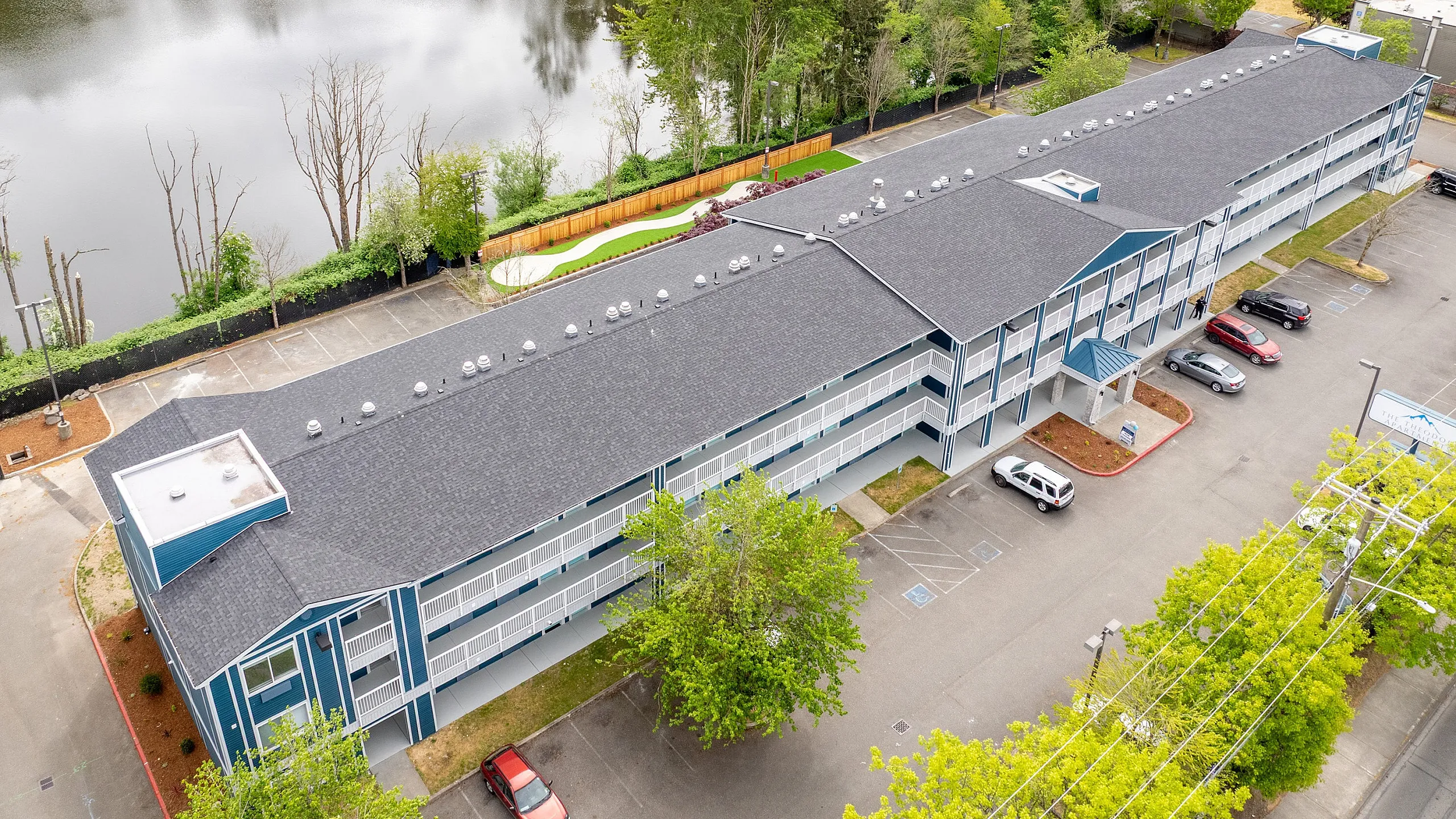 Aerial view of a modern apartment building surrounded by lush greenery and a serene pond, offering a tranquil living environment with ample parking space.