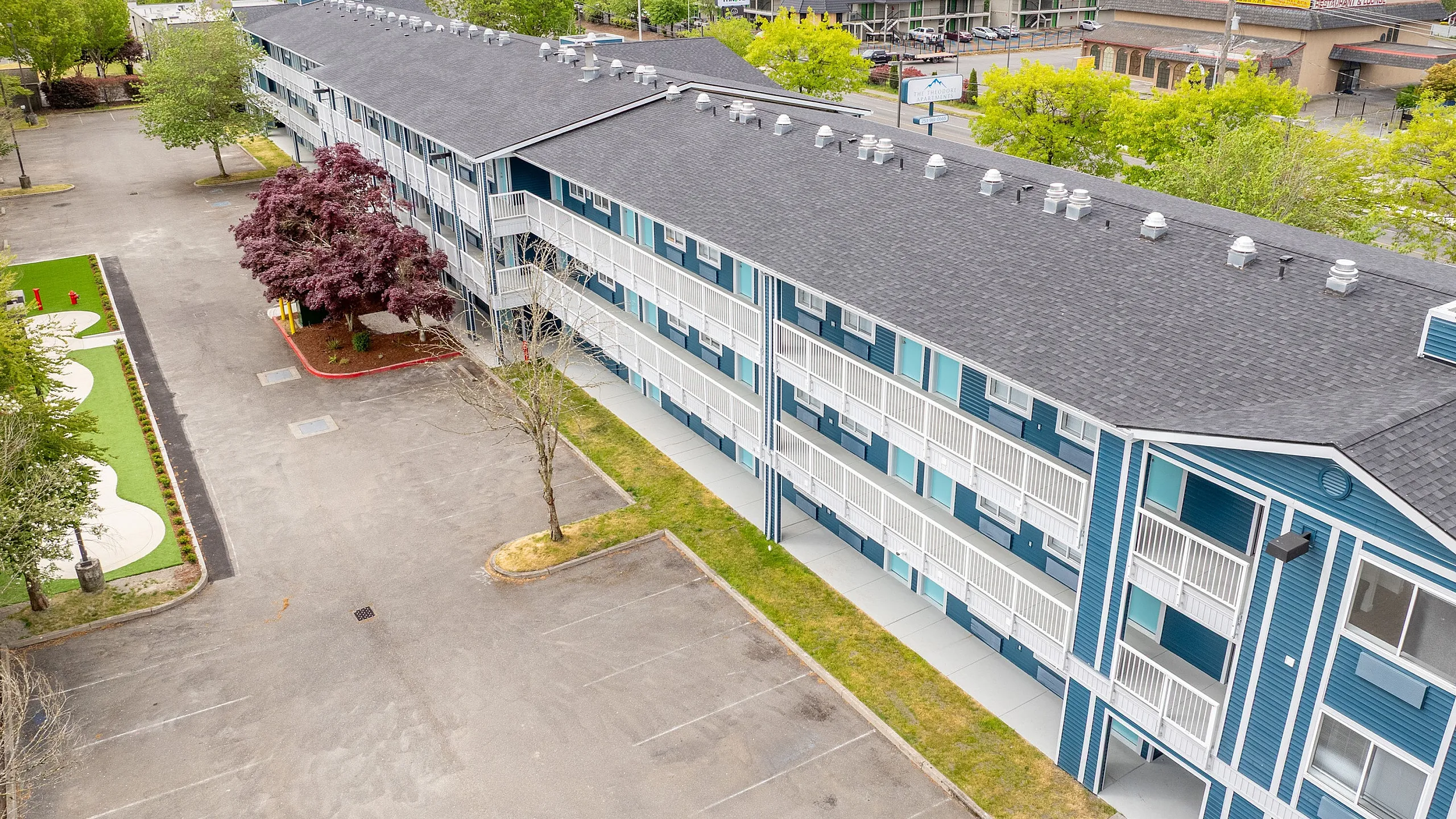 Aerial view of a modern blue-colored apartment complex surrounded by lush greenery, showcasing its expansive layout and ample parking space.