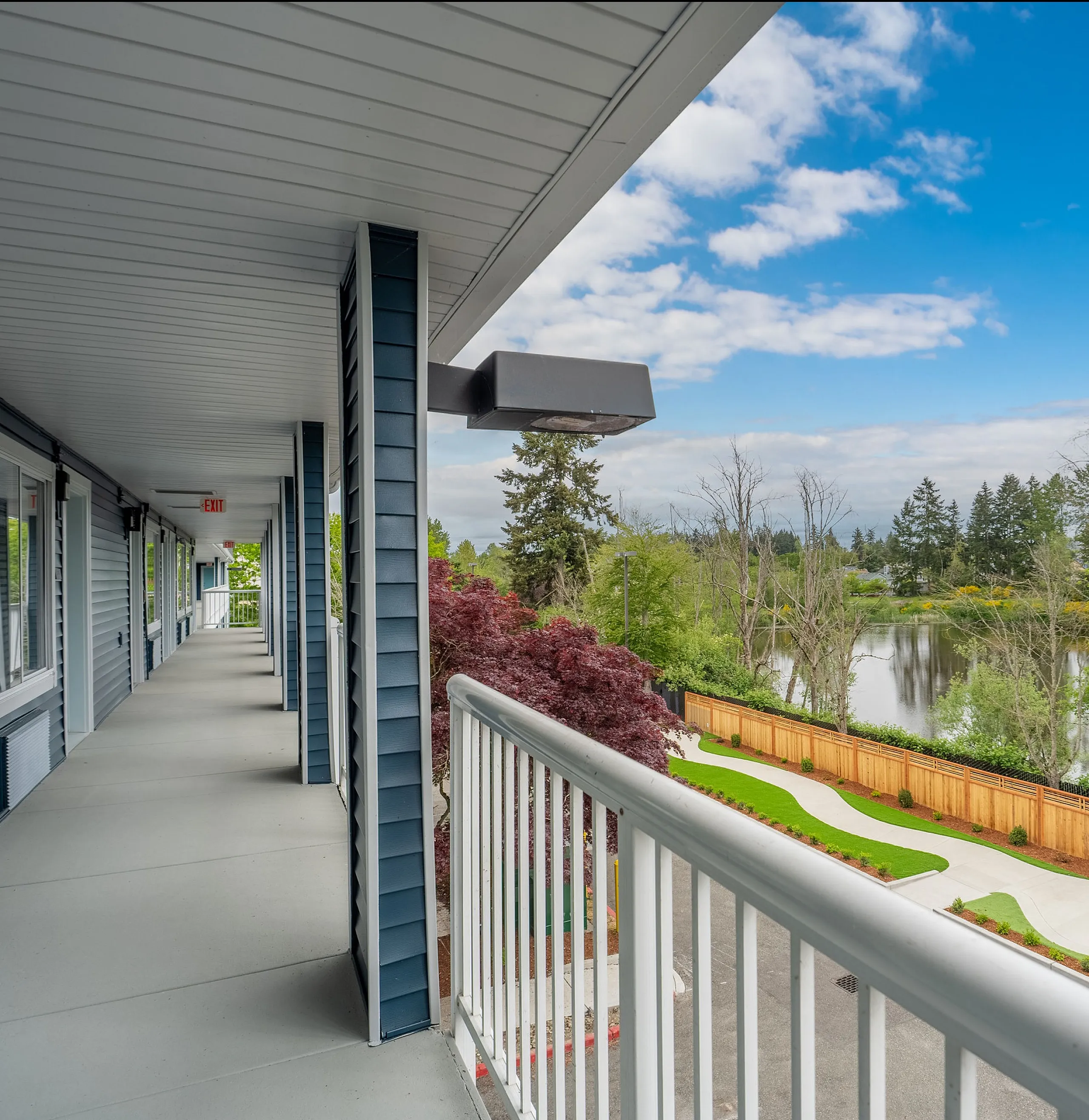 Stunning apartment exterior with a serene lakeside view from a spacious walkway, surrounded by lush greenery and vibrant landscaping.
