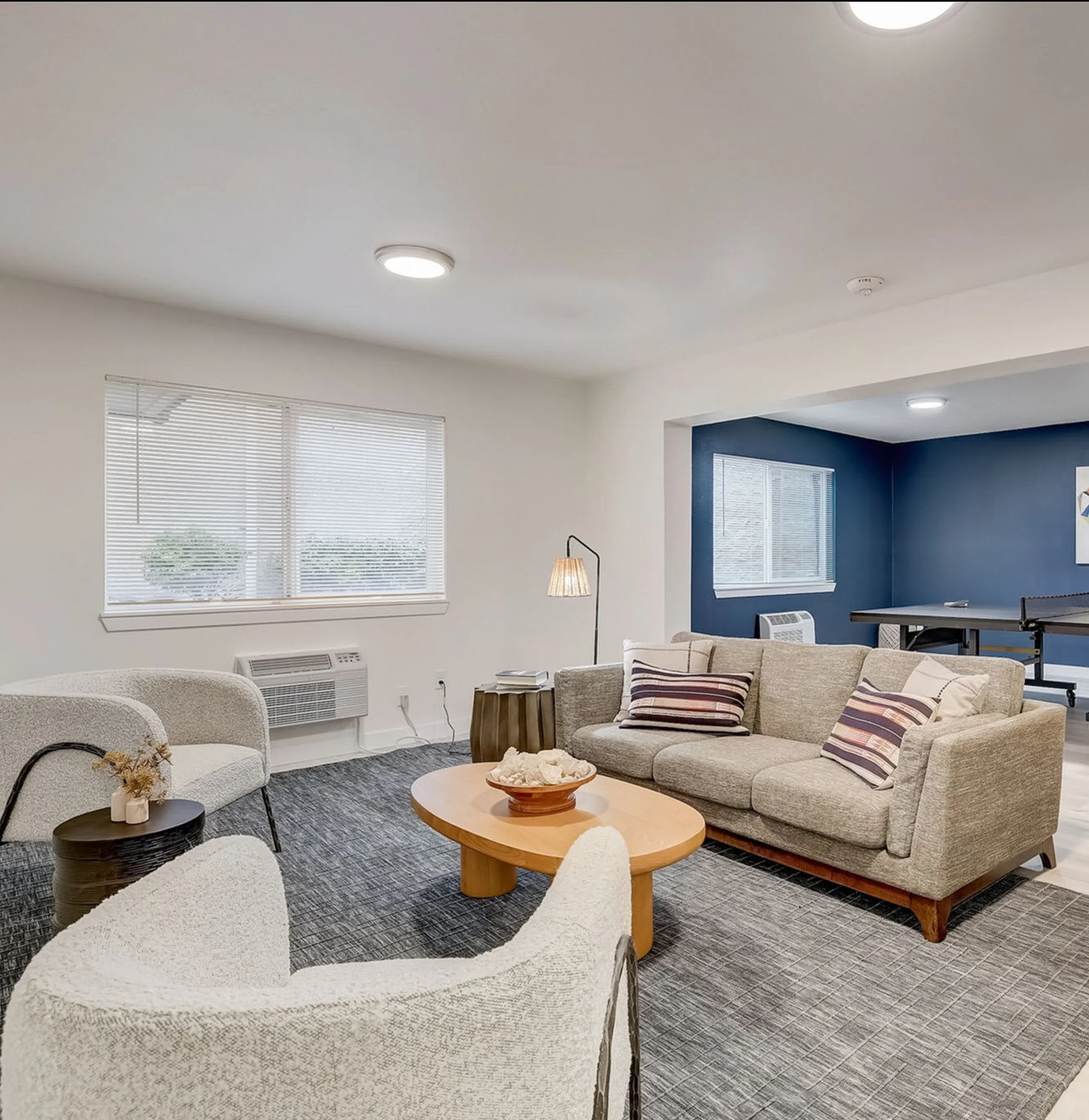 Bright and modern living room featuring a cozy seating area, stylish gray sofa, and chic bar stools, with a vibrant ping-pong table nook in a tastefully decorated open layout.