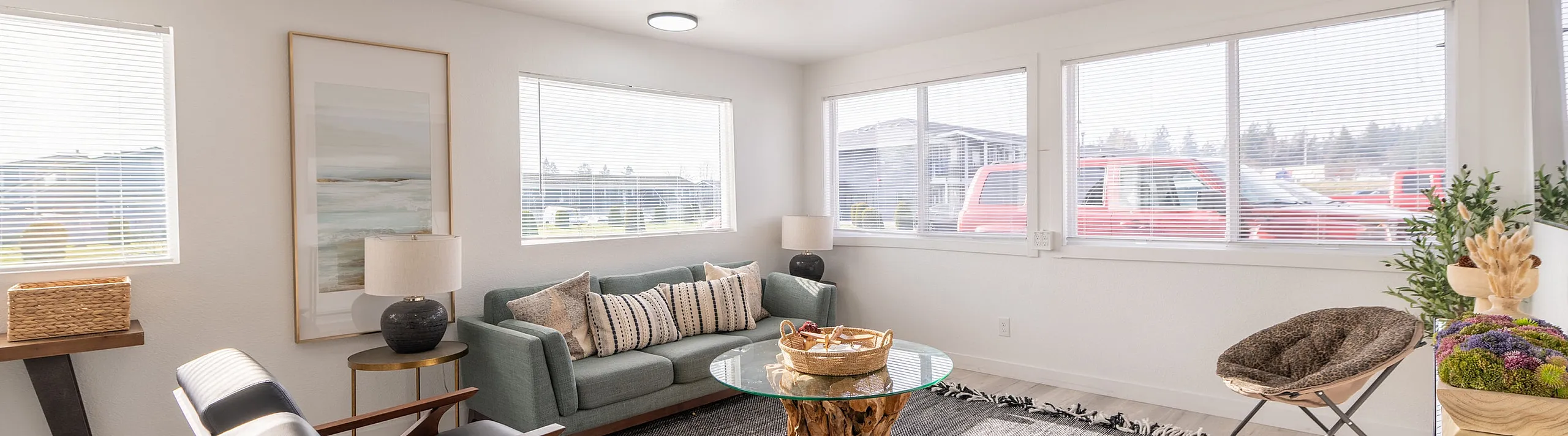 Bright living room with modern decor, featuring large windows, a stylish green sofa, accent chairs, and a unique glass coffee table.