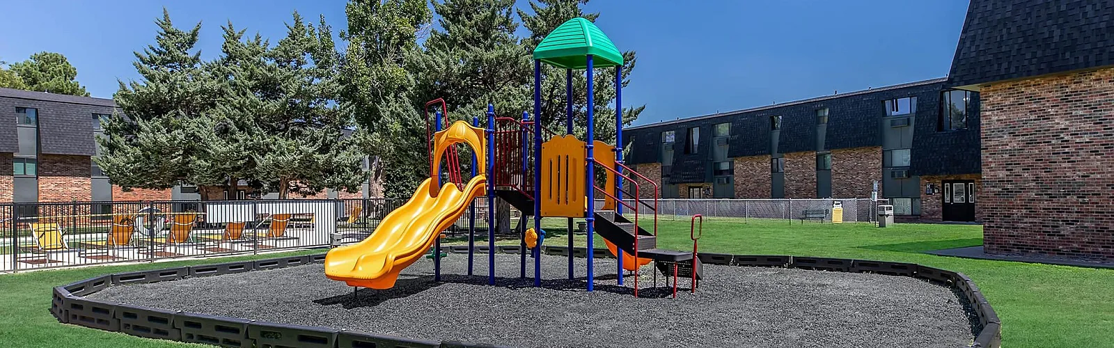 Vibrant playground with colorful slides and climbing structures in a serene apartment complex setting, surrounded by lush green lawns and modern brick buildings.