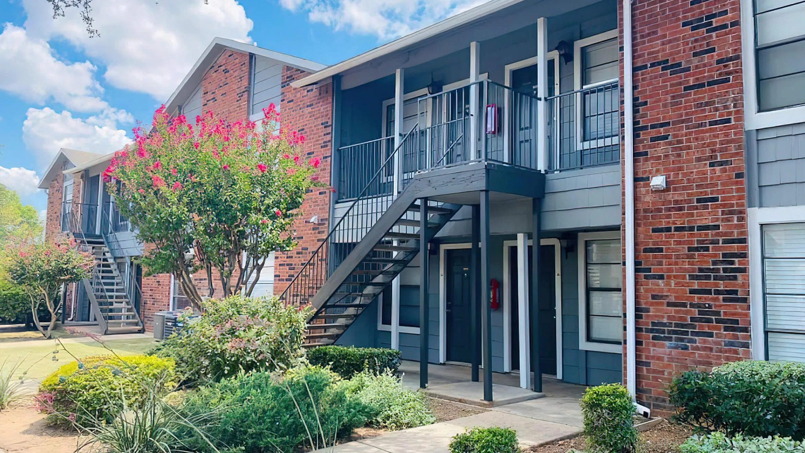 Charming brick apartment complex with lush landscaping and vibrant flowering shrubs under a bright blue sky.