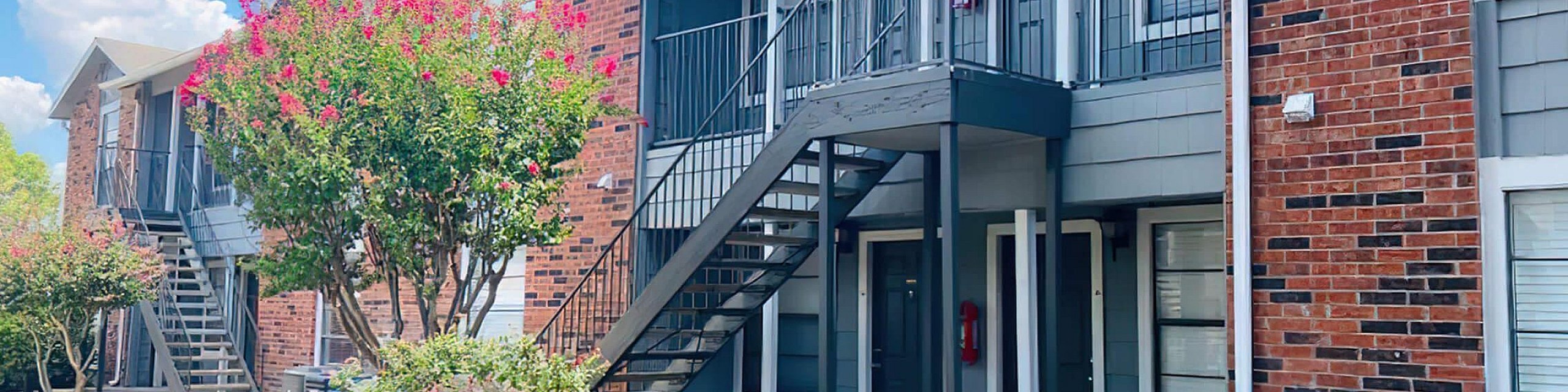 Charming brick apartment building with inviting staircases, surrounded by lush landscaping and vibrant flowers under a sunny blue sky.