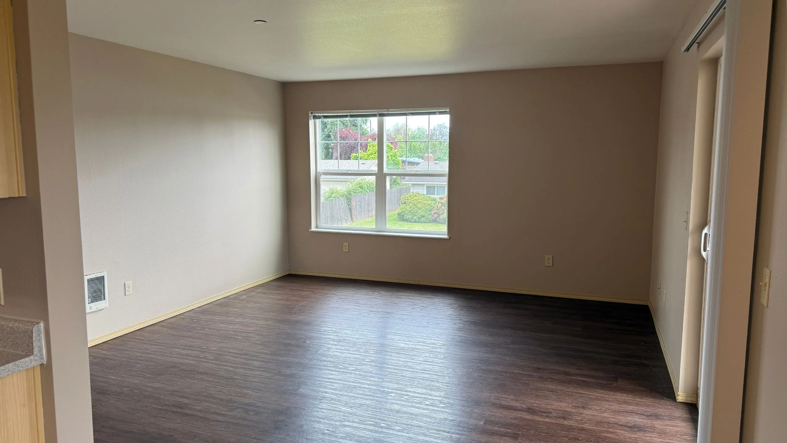 Bright and airy living room with large window, offering natural light and a view of lush greenery, complemented by elegant dark wood flooring.