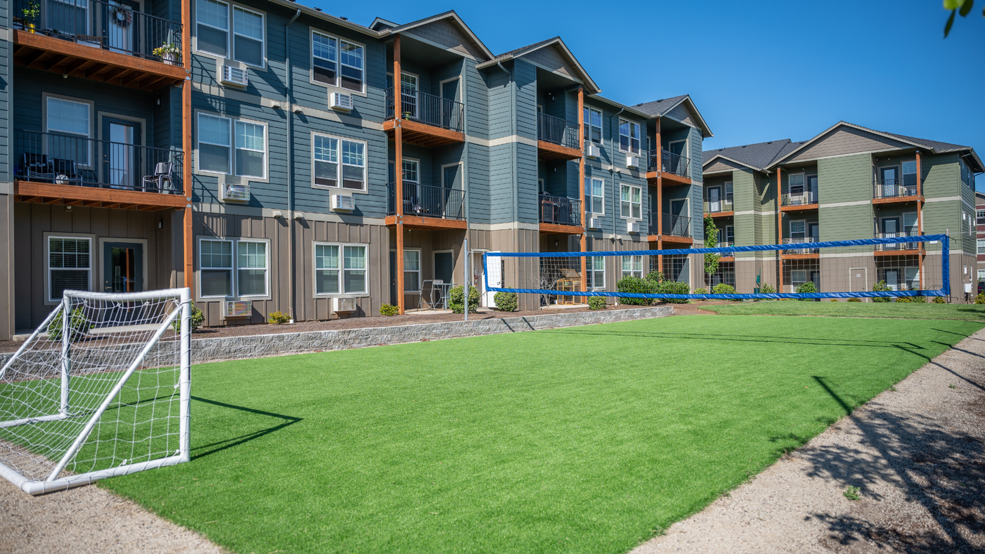 Modern apartment complex featuring vibrant green lawn with soccer goal and volleyball net, perfect for outdoor activities and community living.