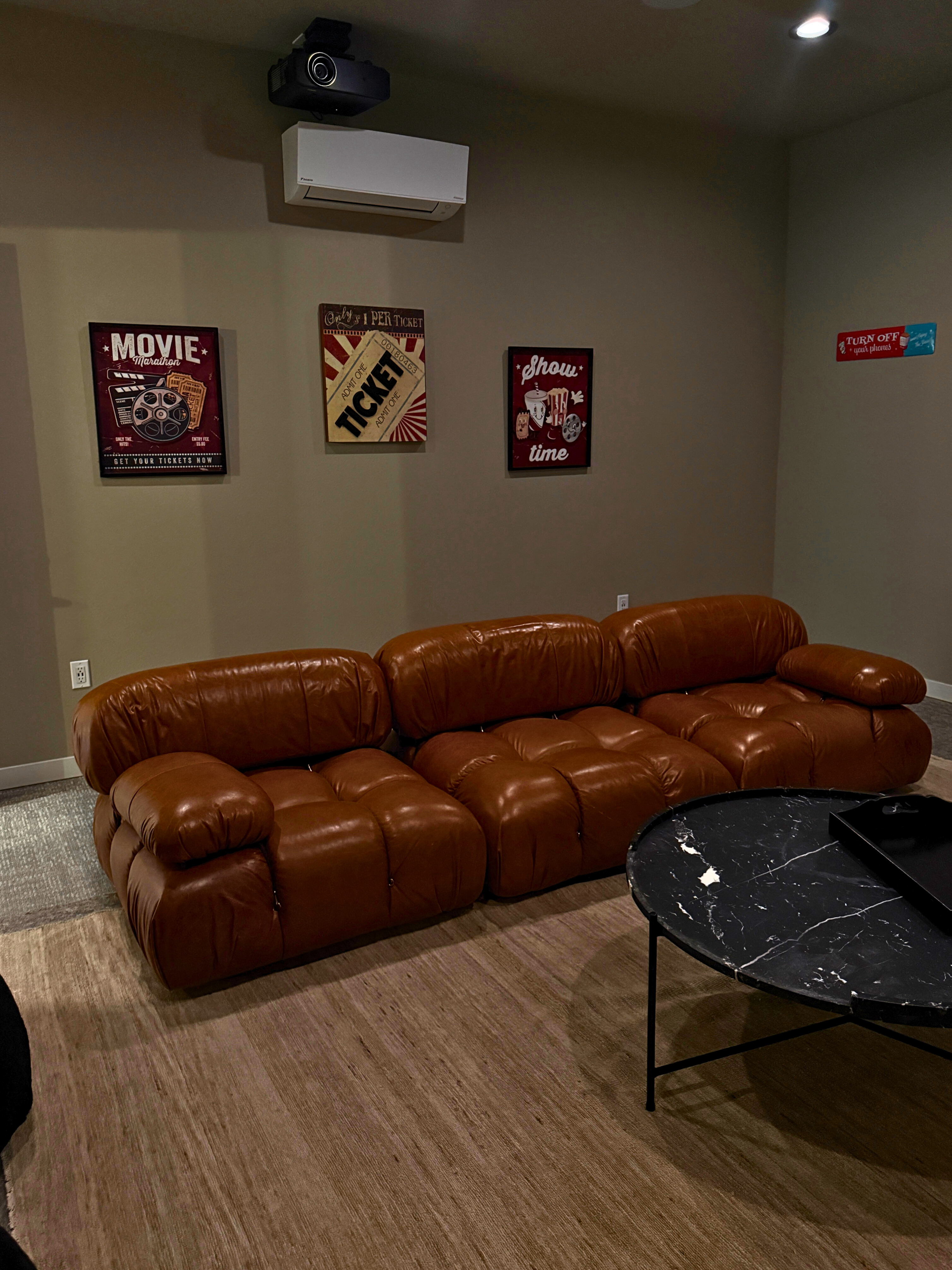 Cozy home theater room with plush brown leather seating, a sleek black marble coffee table, and vintage-style movie posters on the walls.