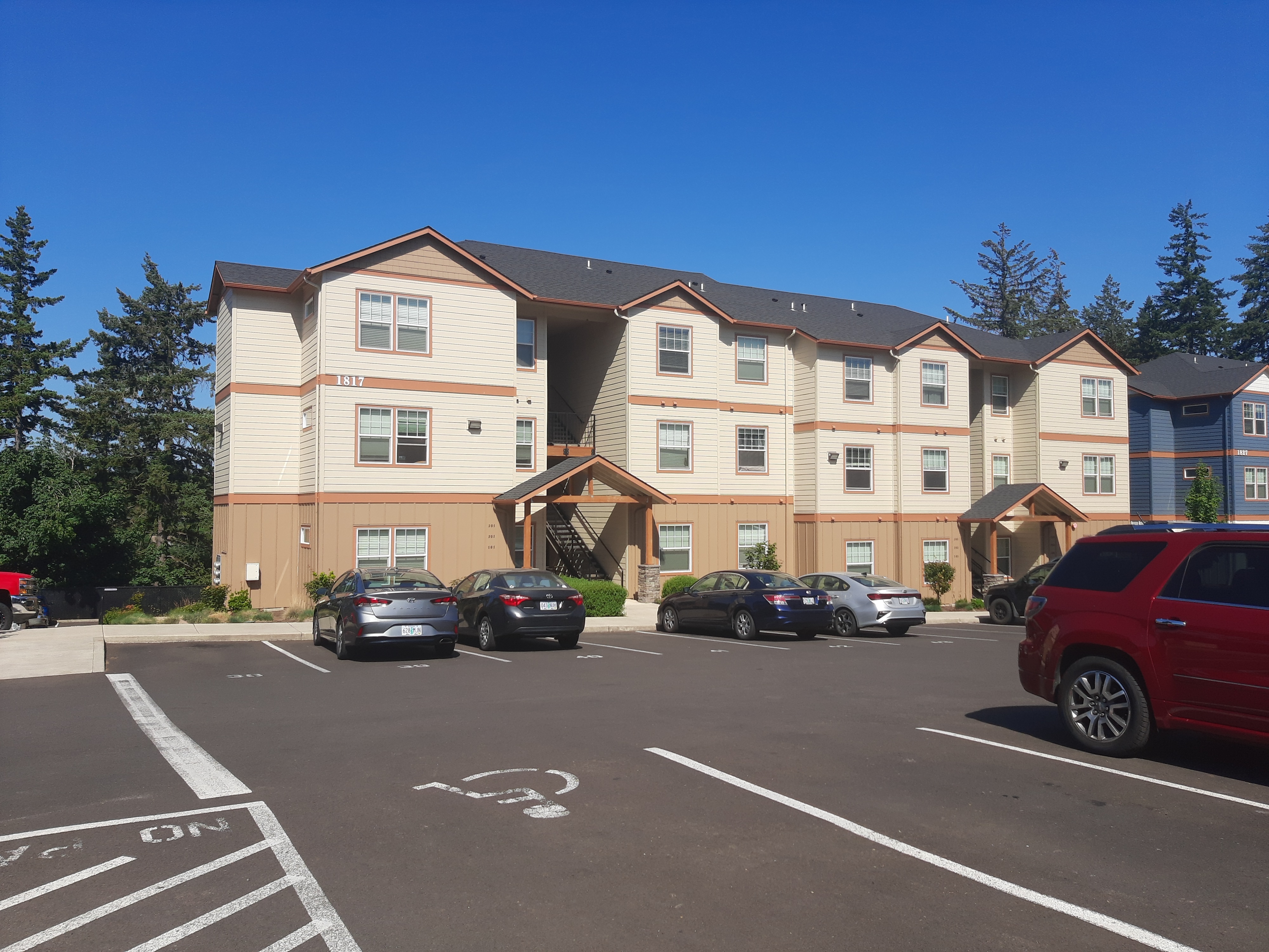 Charming three-story apartment building with modern design, ample parking, and lush greenery under a clear blue sky.