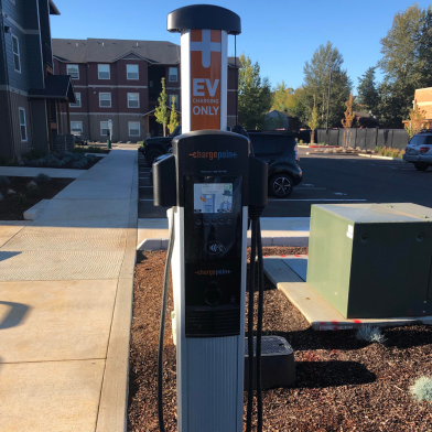 Modern EV charging station in a sunny parking area, surrounded by lush landscaping and contemporary apartment buildings.