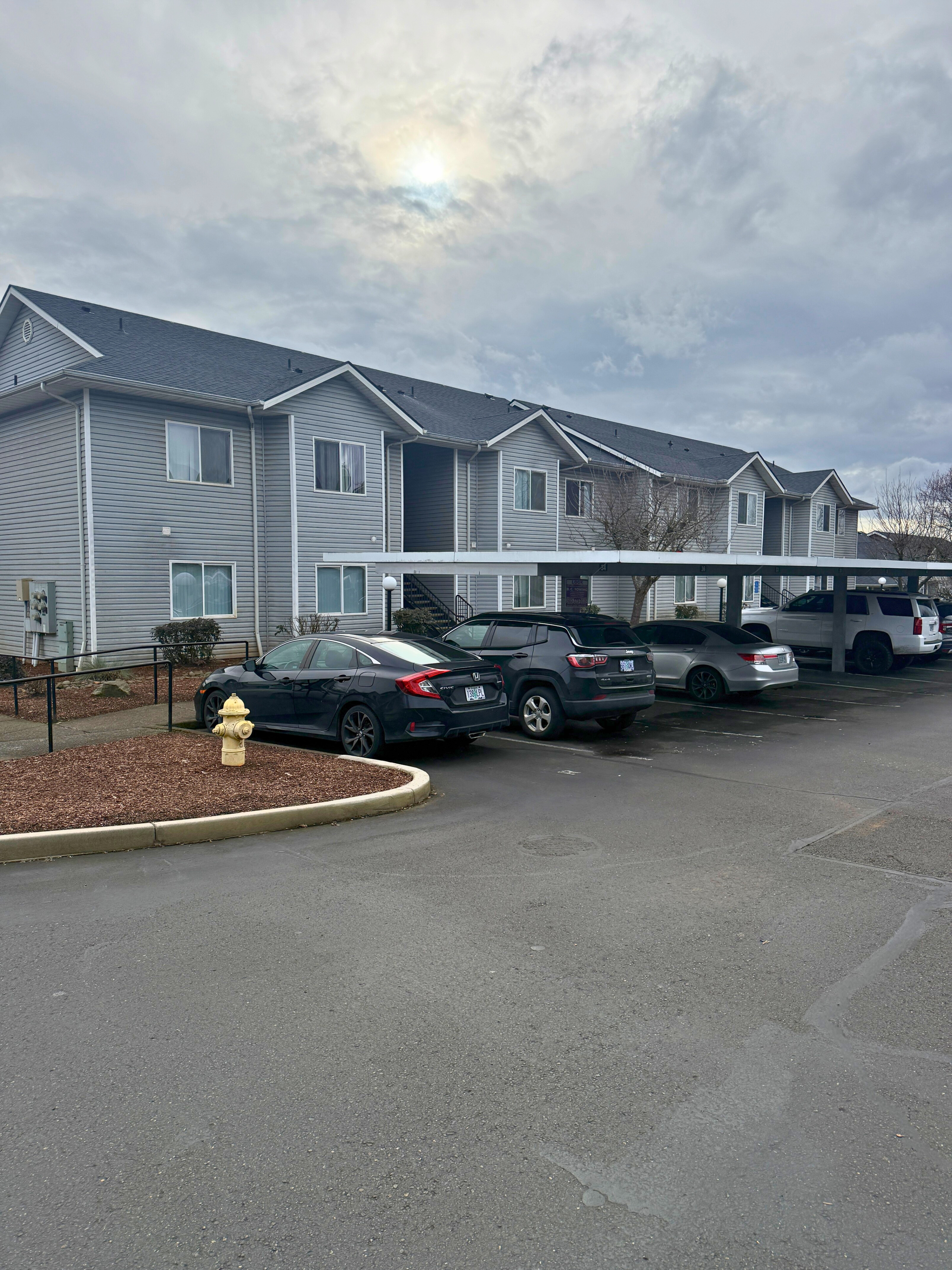 Charming two-story apartment building with modern gray siding, ample parking, and landscaped surroundings under a partly cloudy sky.