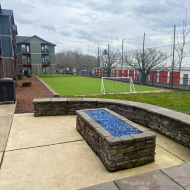 Inviting outdoor amenity area with a blue glass fire pit, lush green lawn, and modern apartment buildings in the background.