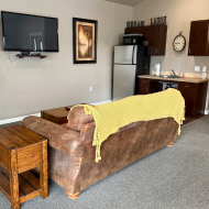 Cozy living space featuring a plush brown sofa with a yellow throw, modern kitchenette, and a wall-mounted TV for ultimate comfort and convenience.