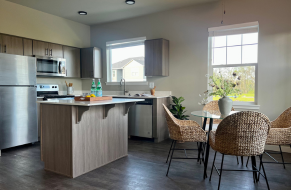 Modern kitchen with stainless steel appliances, wooden cabinets, and a cozy dining area with woven chairs and natural light.