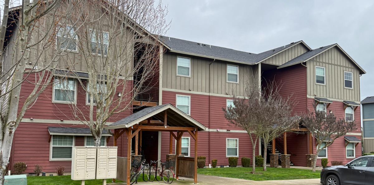 Charming three-story apartment building with rustic red and taupe exterior, featuring a cozy porch and lush greenery.