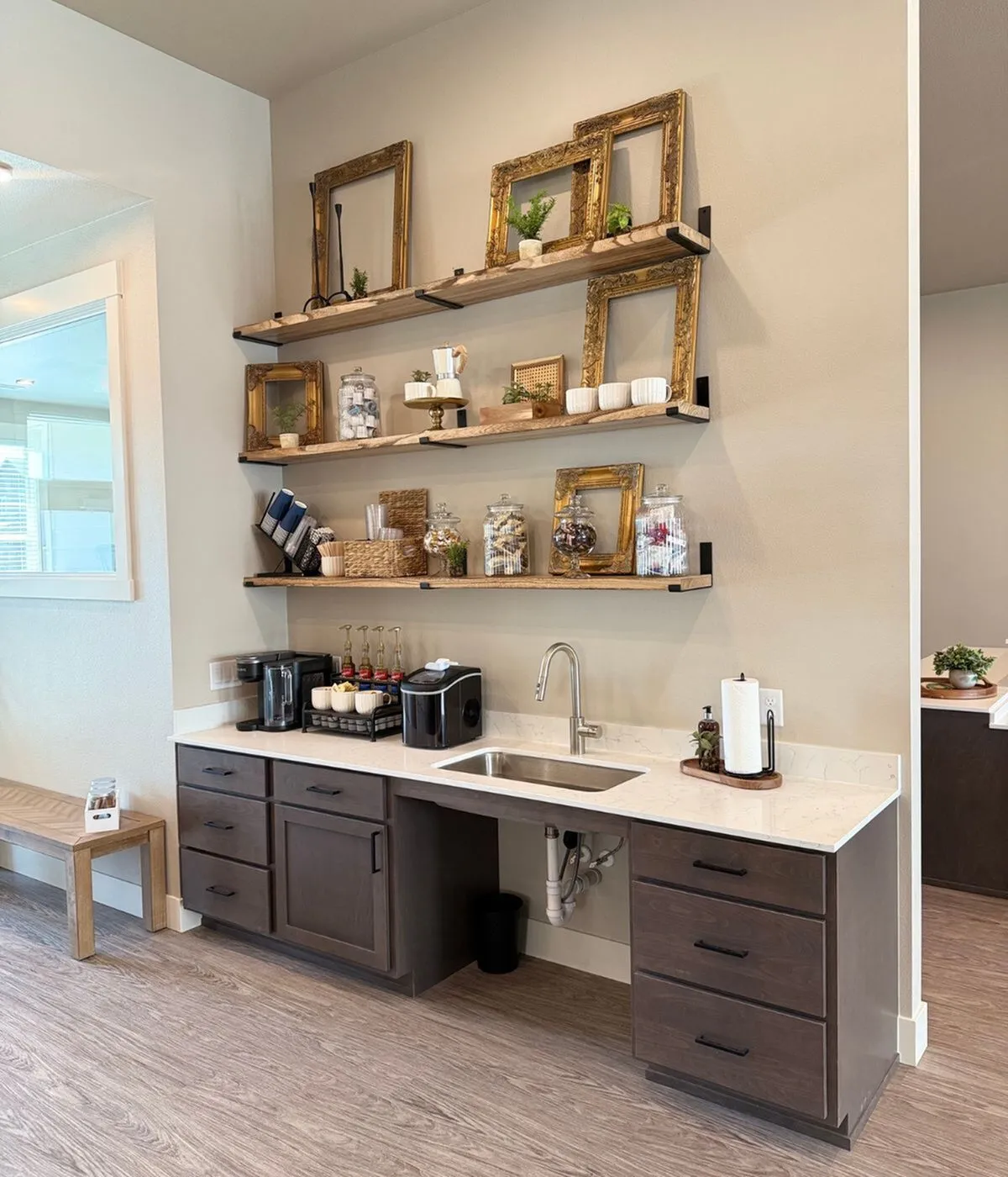 Elegant kitchen nook with chic wooden shelves displaying picture frames and plants, complemented by a modern coffee bar and sleek under-sink storage, creating a stylish and functional space.