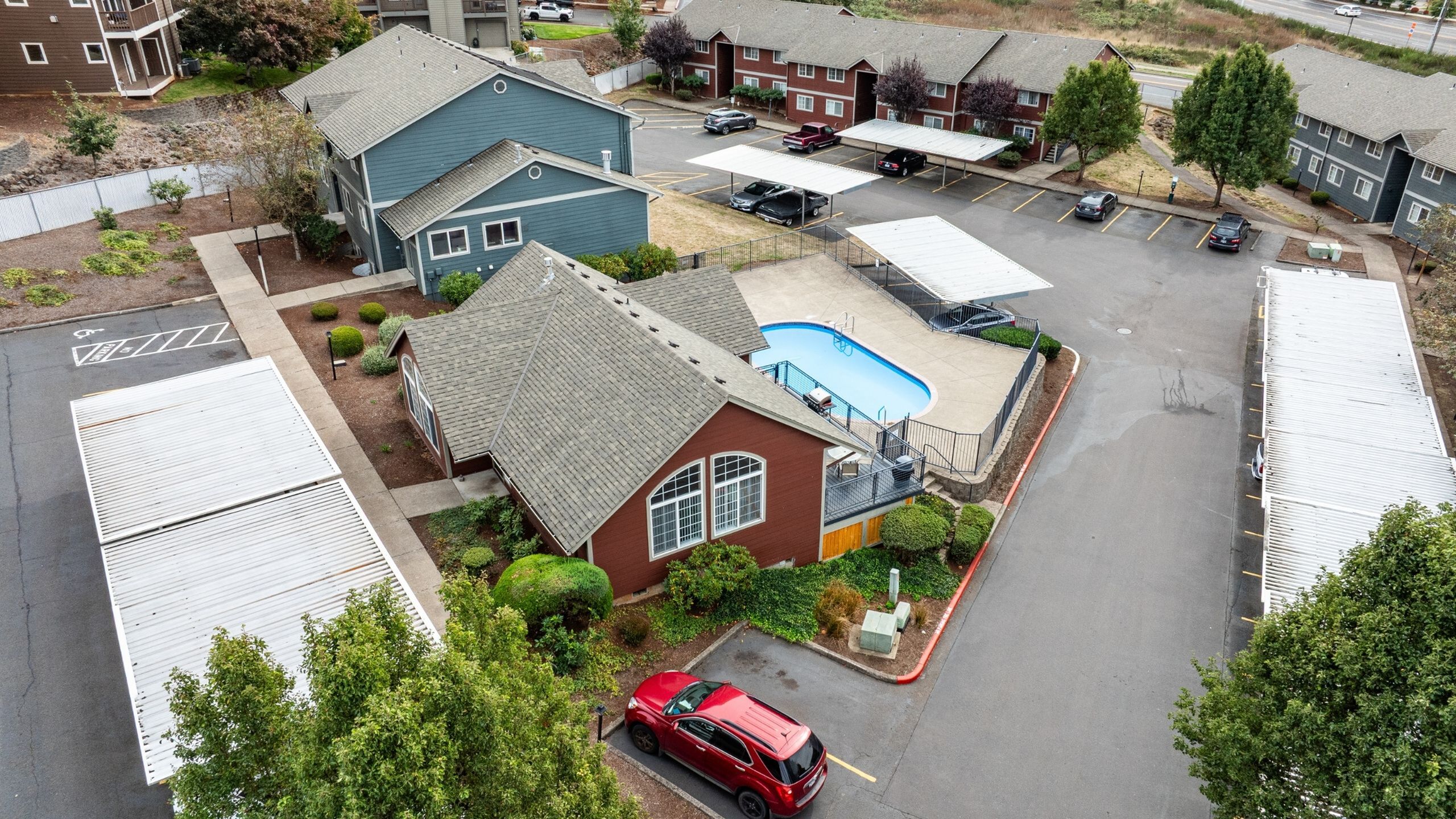 Charming aerial view of a residential complex featuring neat grey and red buildings, a glistening blue swimming pool, and ample parking area surrounded by lush greenery.