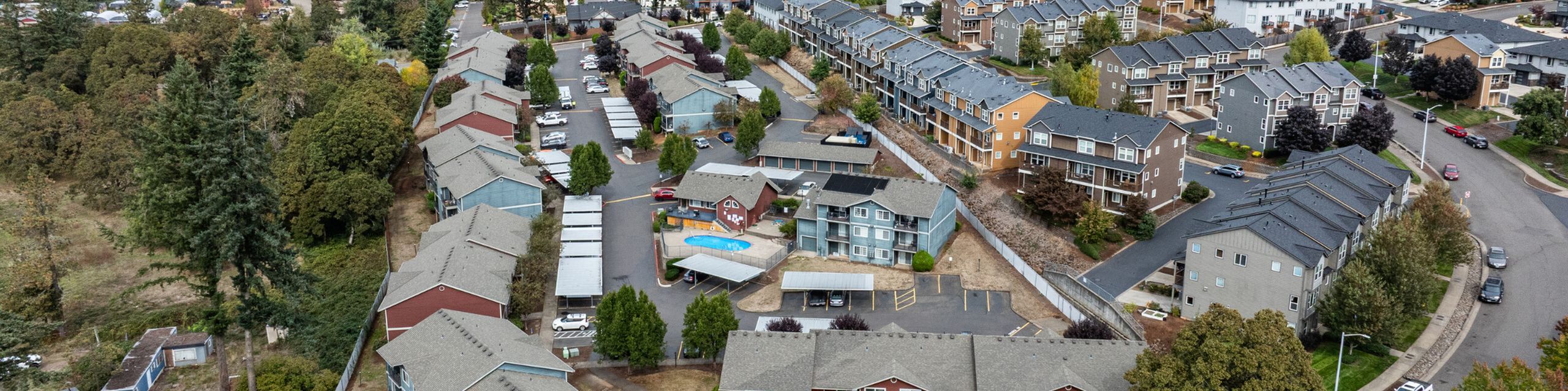 Aerial view of a charming residential complex with multiple modern apartment buildings surrounded by lush greenery, featuring a central swimming pool and ample parking spaces.