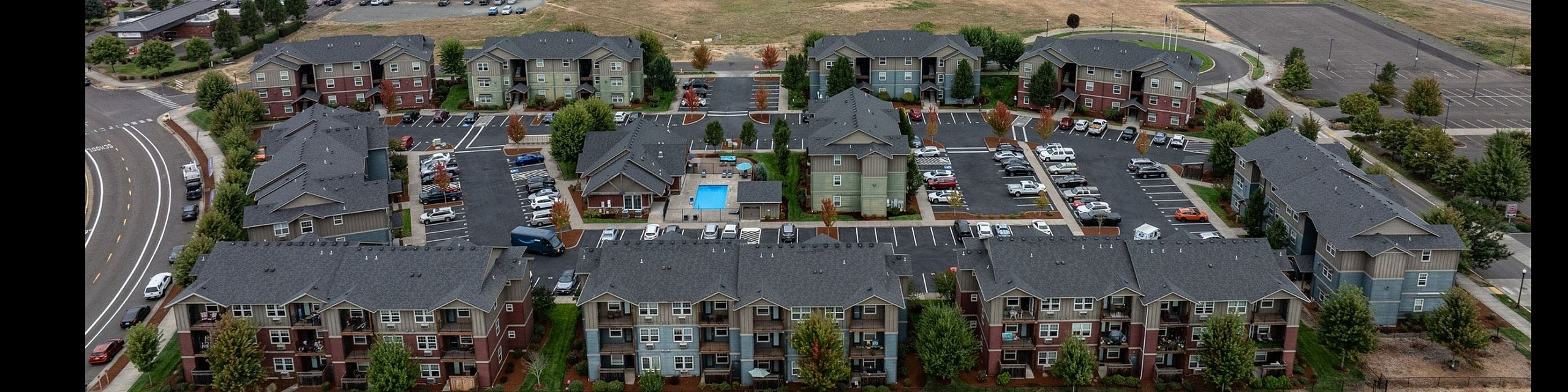 Aerial view of modern apartment complex surrounded by lush greenery and scenic countryside, offering tranquil living with a communal pool and ample parking space.
