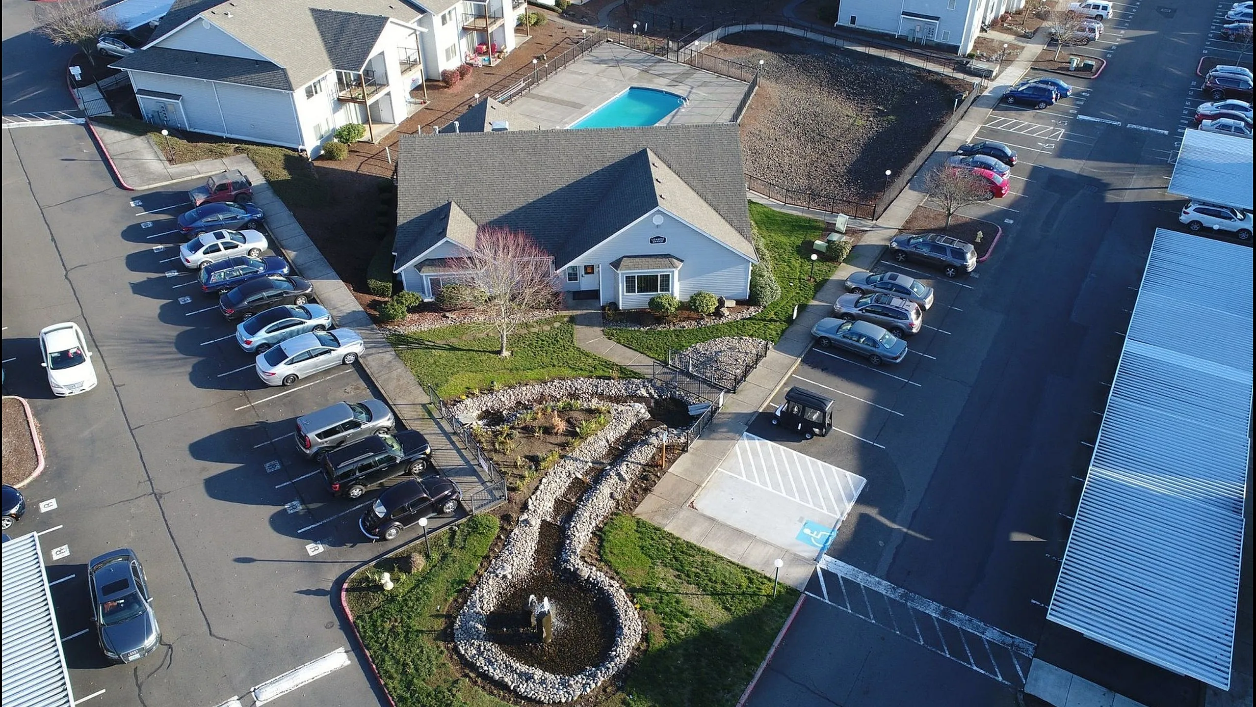 Aerial view of a charming apartment complex featuring a beautiful landscape with a fountain, ample parking spaces, and a pristine swimming pool.
