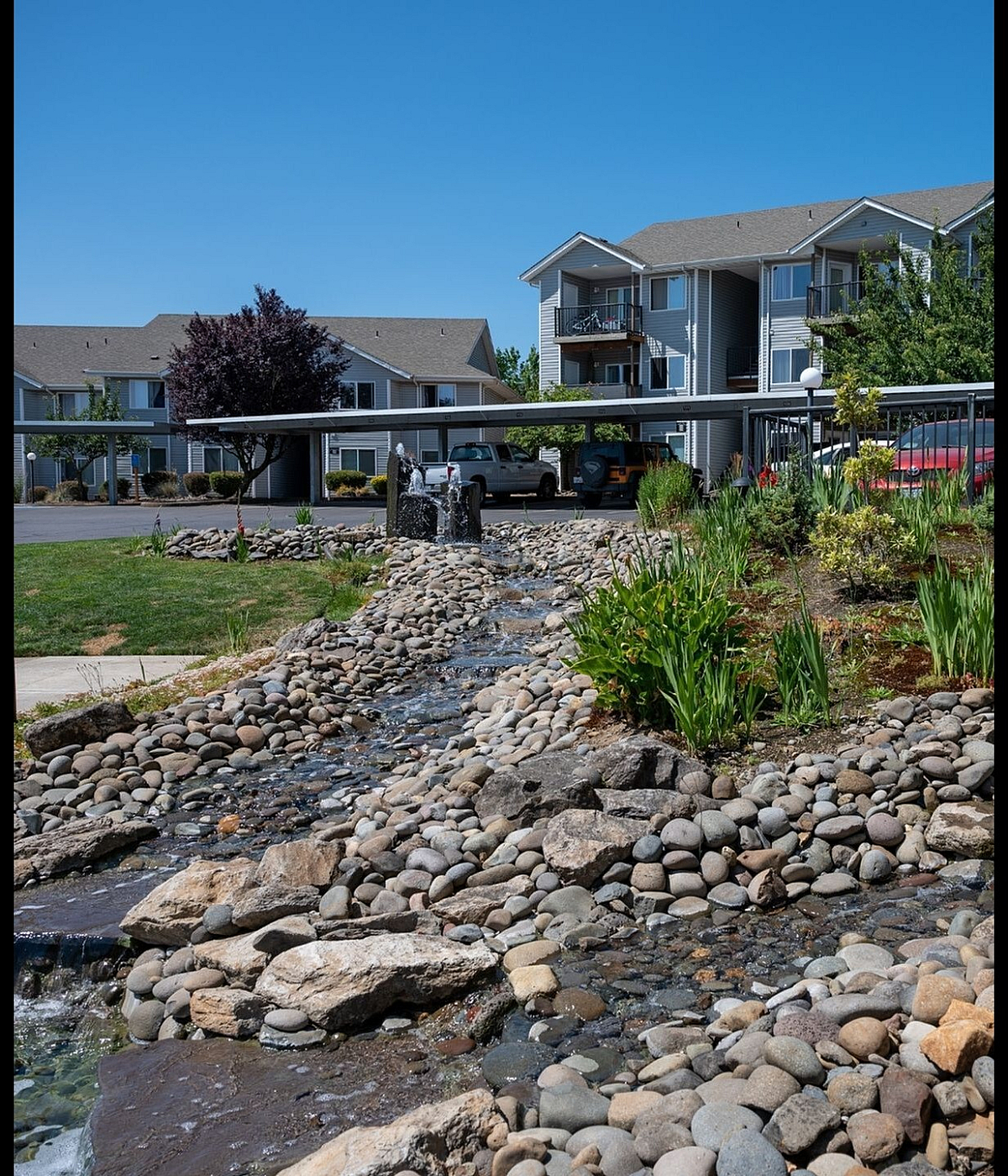 Charming apartment complex with manicured landscapes and a serene rock-lined water feature under clear blue skies.