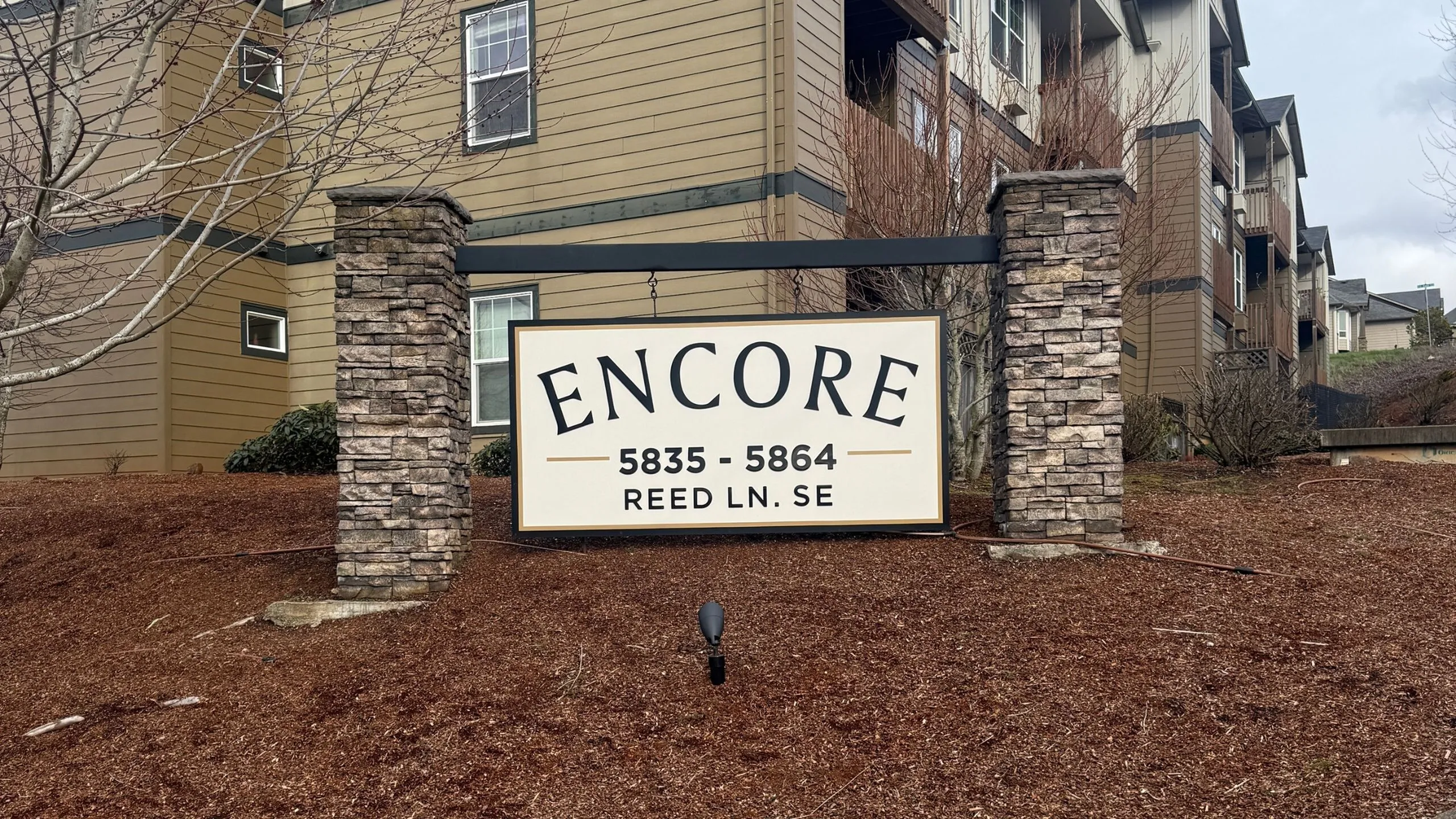 Charming apartment complex entrance with stone pillars supporting a welcoming Encore sign, surrounded by lush landscaping.