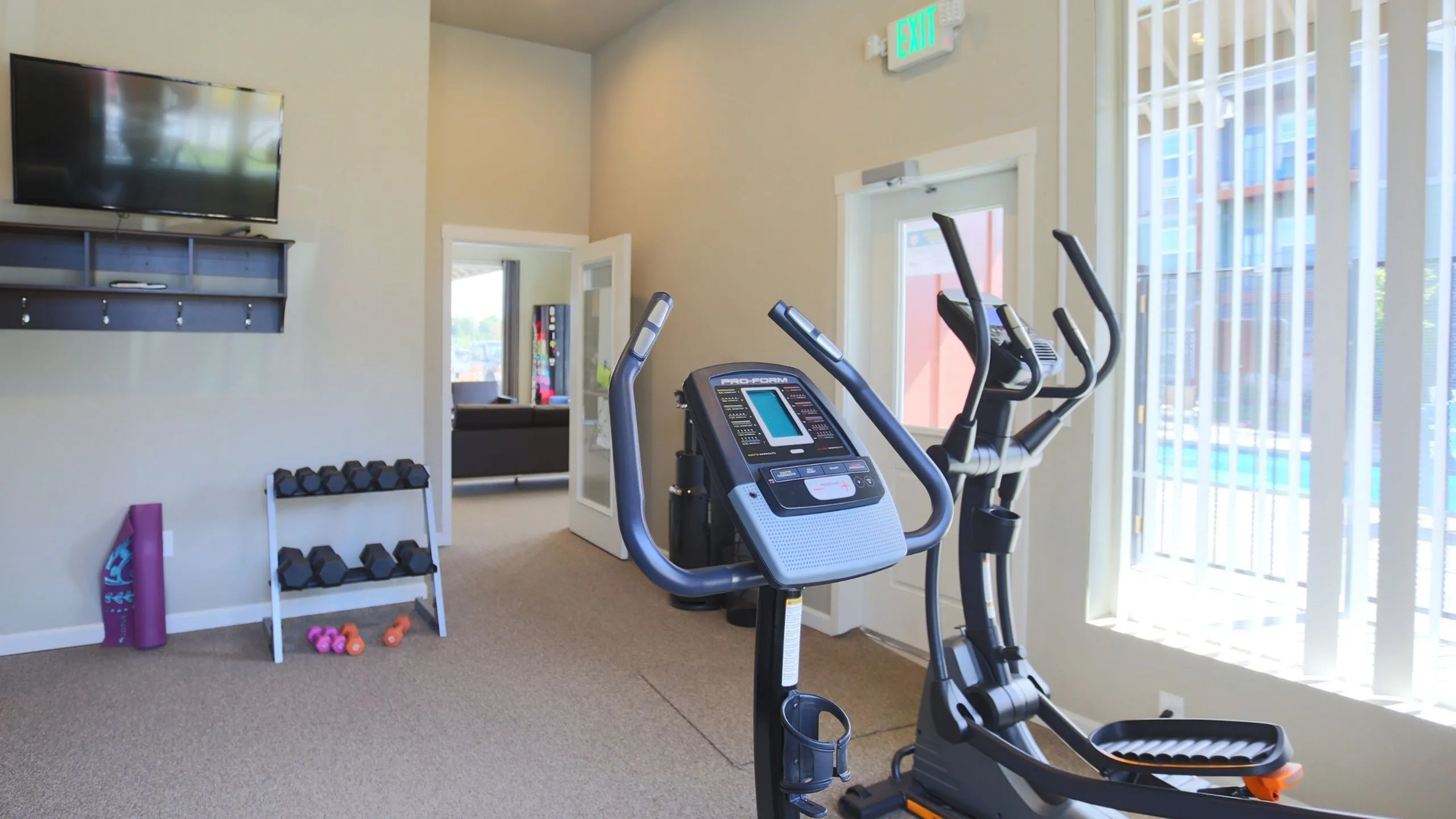 Modern apartment fitness center with elliptical, flat-screen TV, and free weights, offering a bright and inviting workout space.