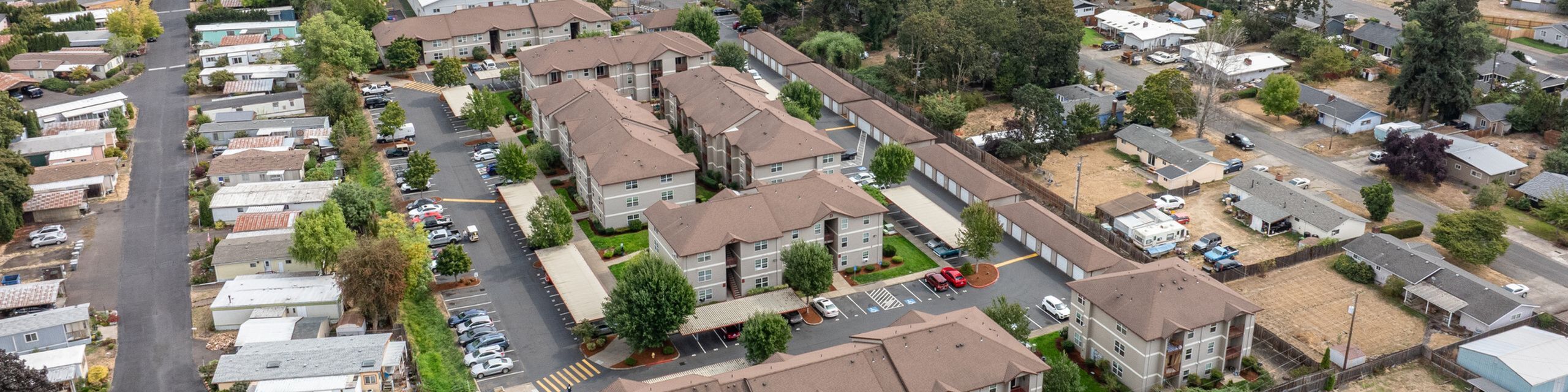 Expansive aerial view of a modern apartment complex surrounded by lush greenery, showcasing ample parking and a well-maintained community setting.