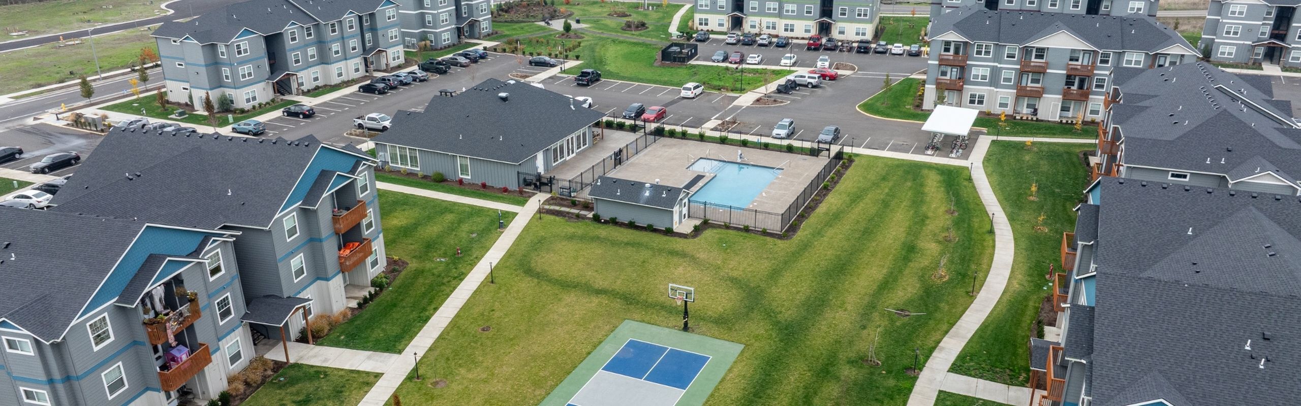 Charming apartment community with vibrant green spaces, a sparkling swimming pool, basketball court, and ample parking, set amidst modern multi-story residential buildings.