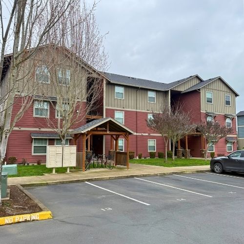 Charming multi-story apartment building with rustic red and beige exterior, surrounded by landscaped greenery and convenient parking.