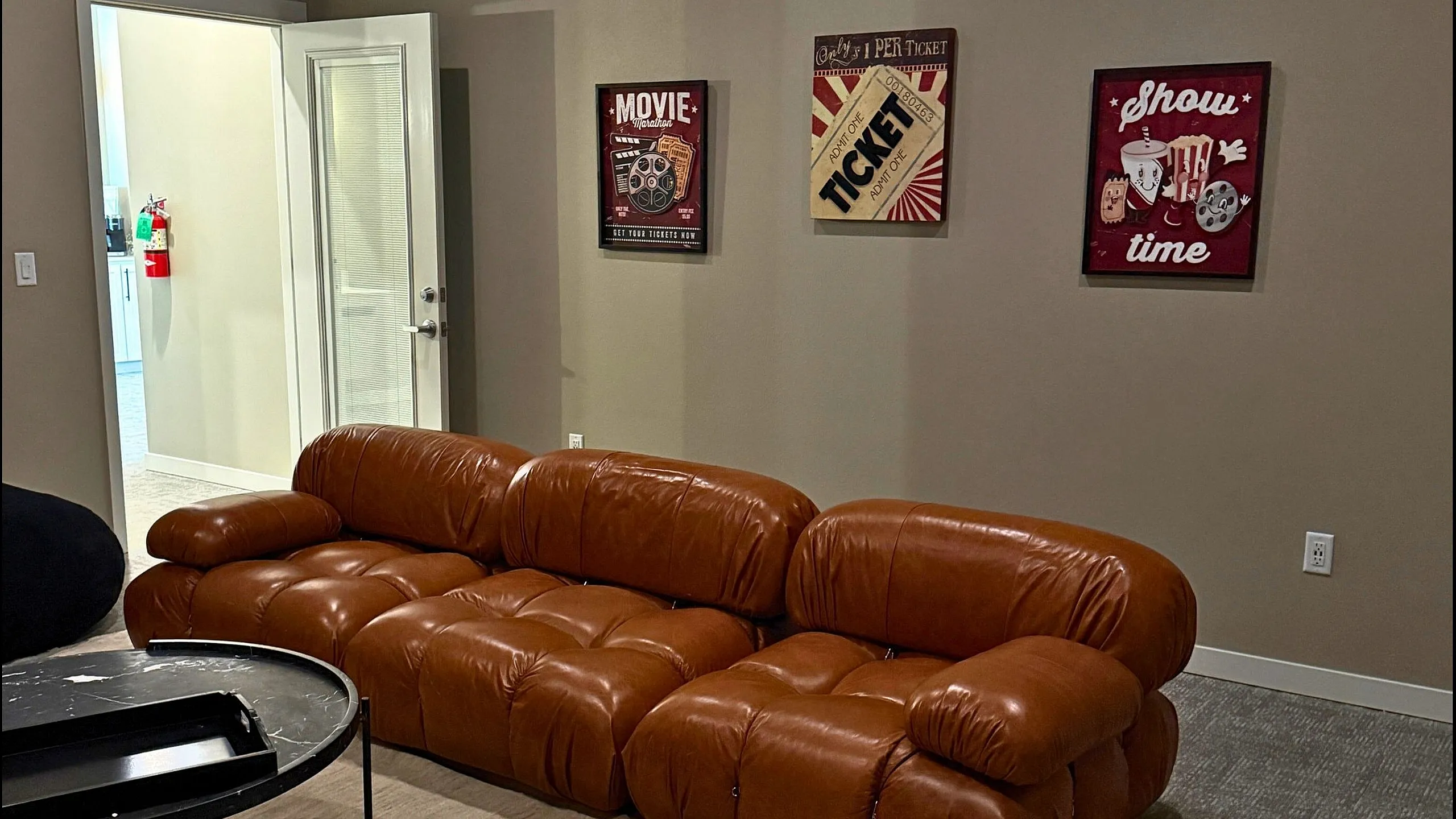 Inviting living space with a cozy brown leather couch, stylish wall art, and an open doorway leading to a bright kitchen.