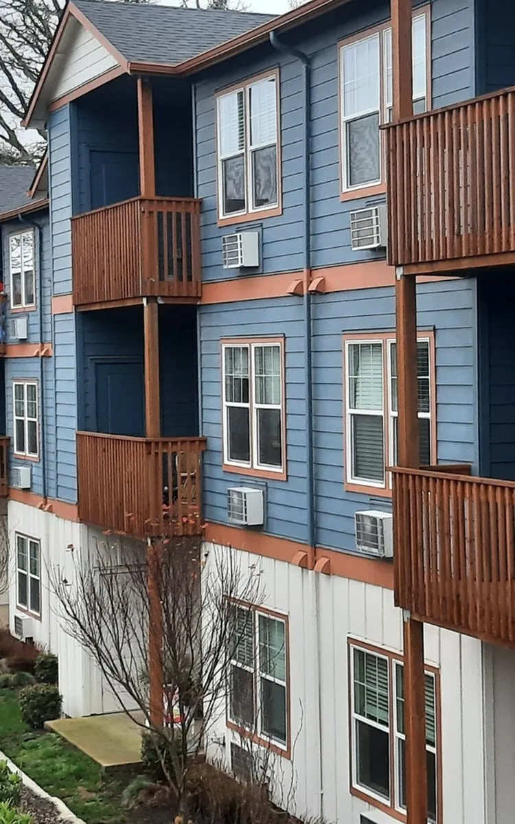 Charming apartment building with blue siding and warm accents, featuring private balconies and lush surroundings.