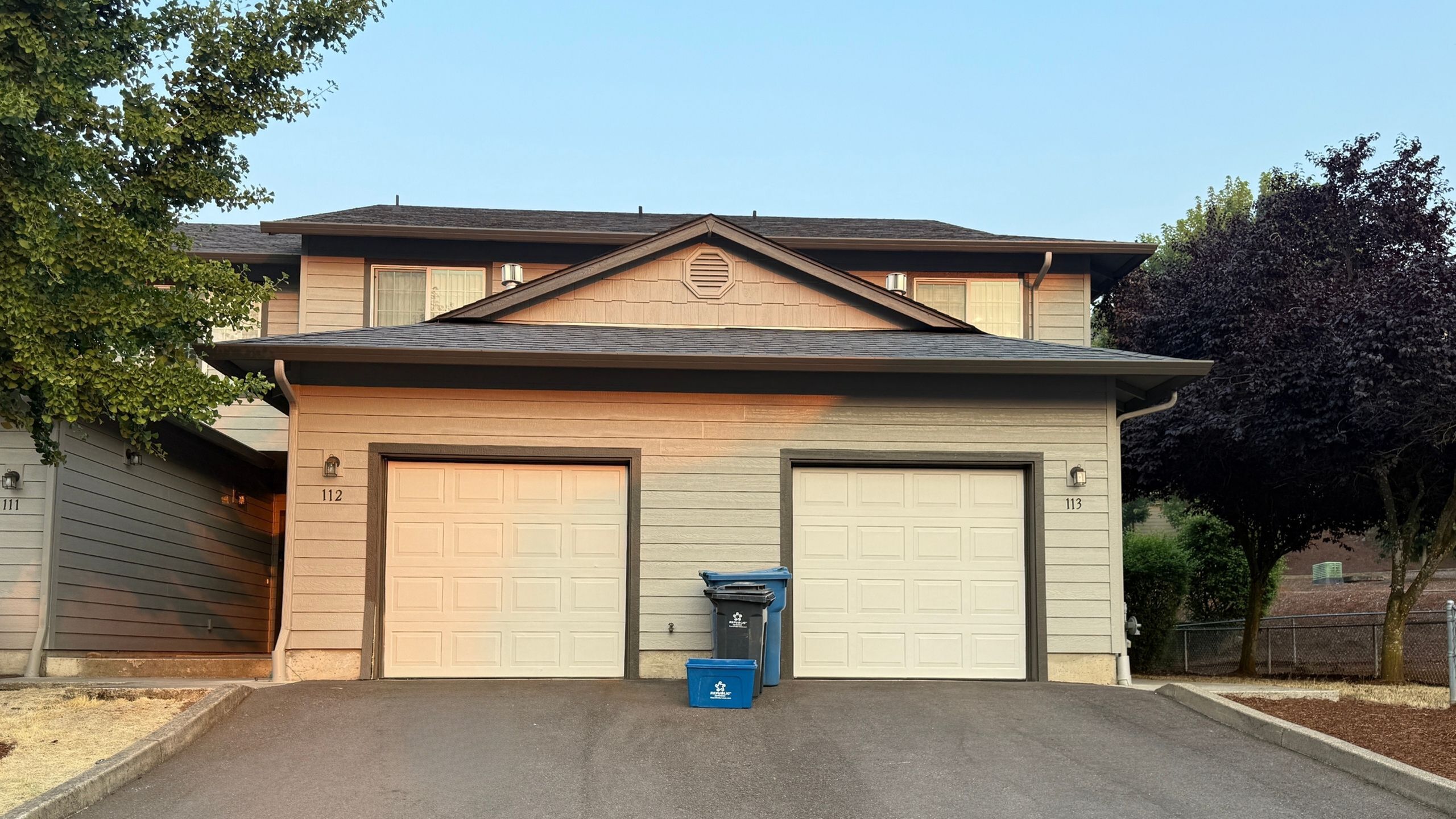 Charming duplex with two garages, surrounded by lush greenery and a well-kept driveway.