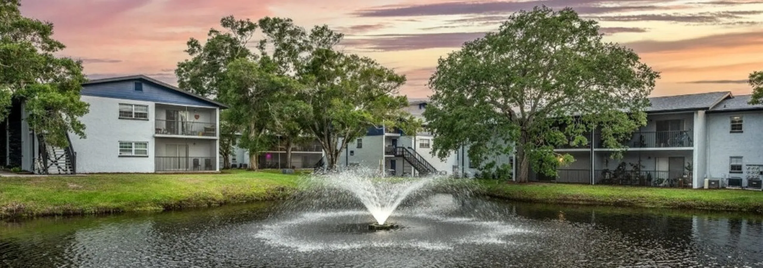 Serene apartment complex with charming pond and fountain at sunset, surrounded by lush greenery and modern buildings.