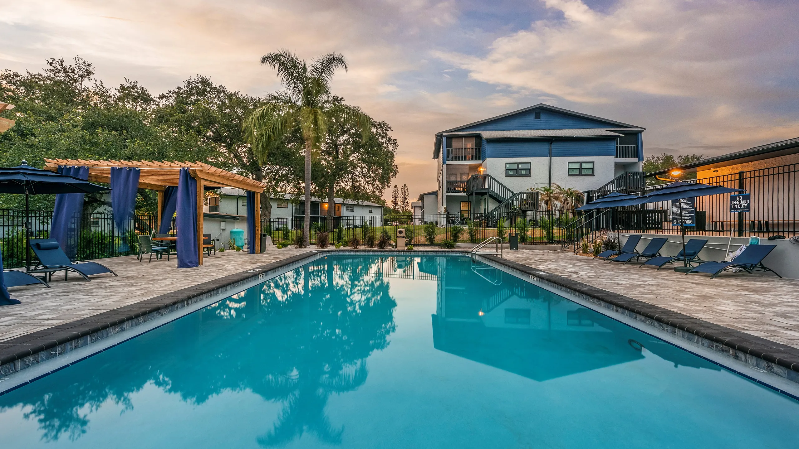 Inviting outdoor pool area with stylish pergola, lounge chairs, and a scenic view of modern apartments at sunset.