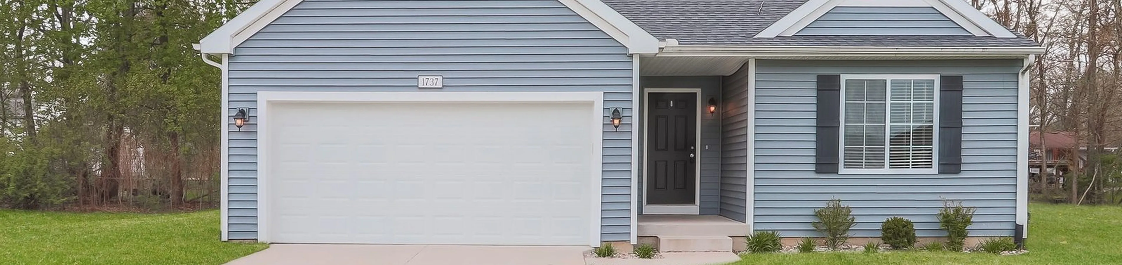 Charming single-story home with blue siding, cozy front porch, and a spacious two-car garage surrounded by lush greenery.