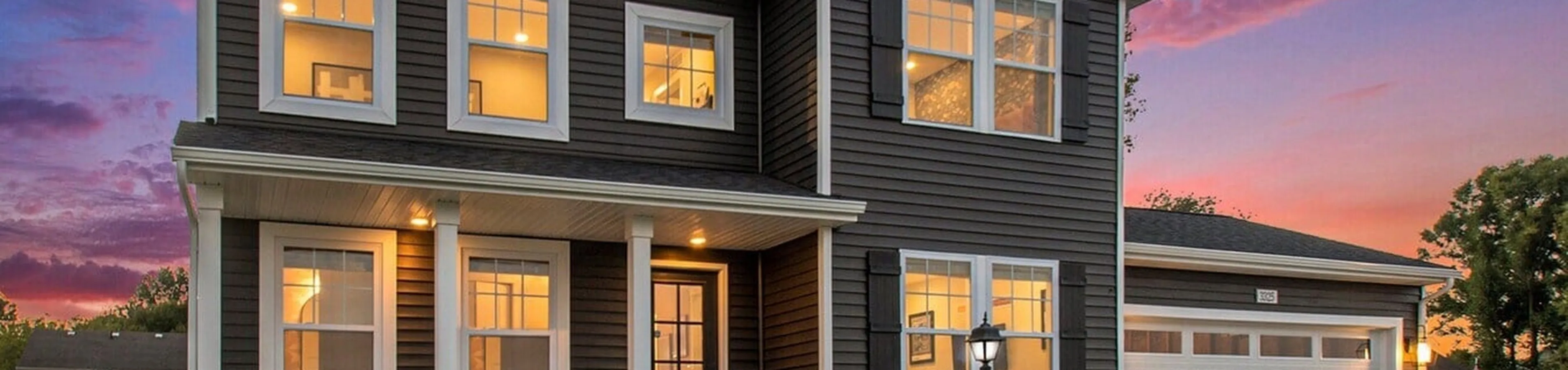 Charming two-story home with classic dark siding and white trim, warmly lit windows, and a welcoming front porch against a vibrant sunset backdrop.