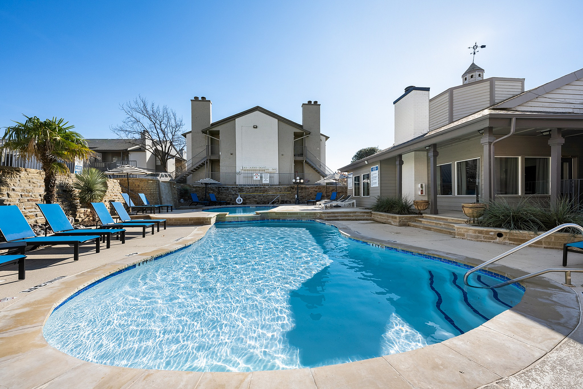 Inviting apartment complex with a sparkling outdoor pool surrounded by modern loungers and lush landscaping under a clear blue sky.