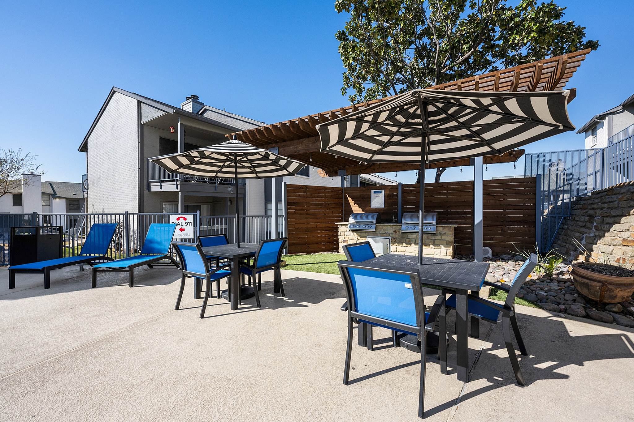Chic outdoor patio with blue chairs and elegant parasols, perfect for relaxation, showcasing modern barbeque grills and stylish fenced outdoor area.