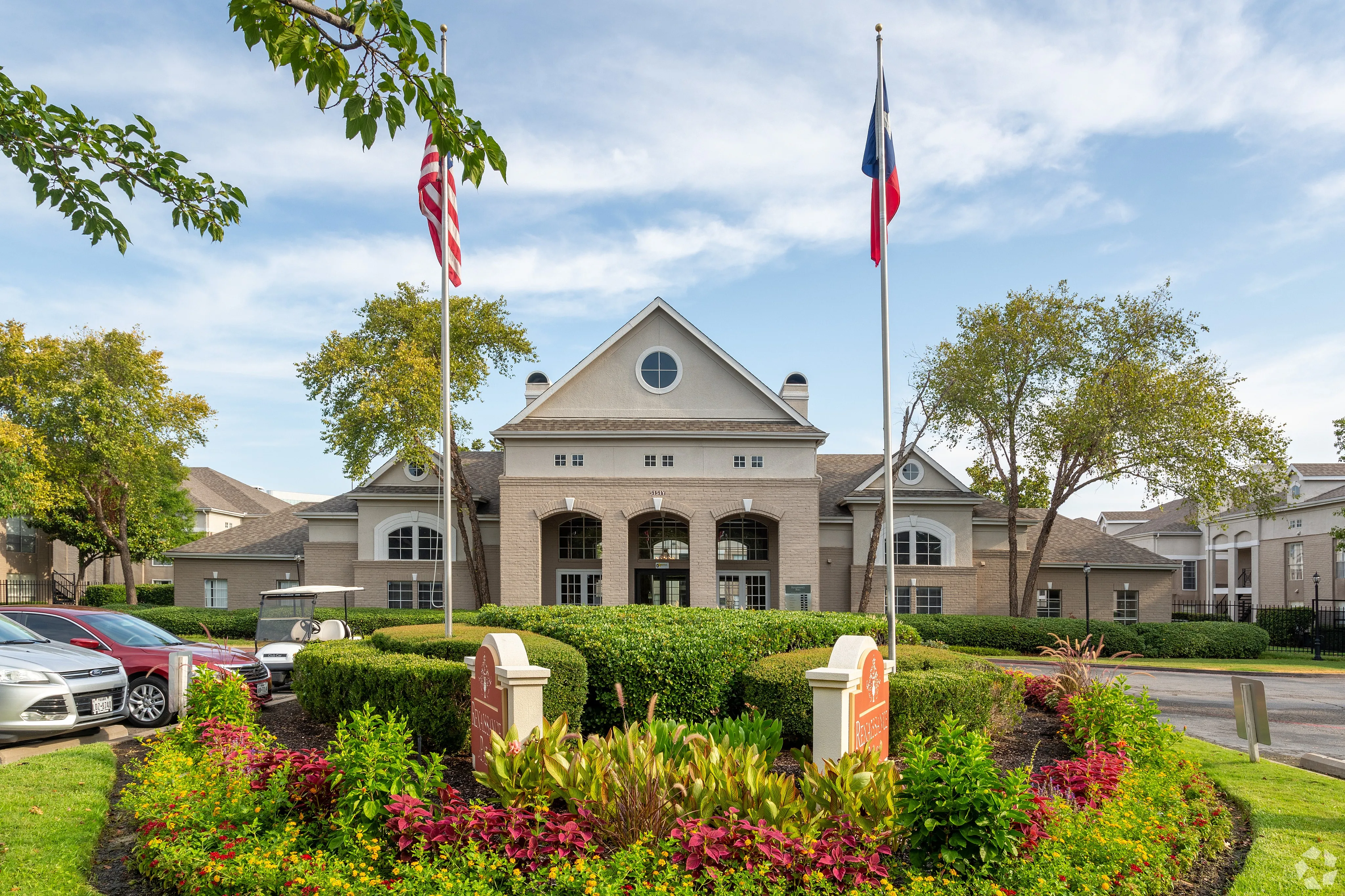 Inviting residential complex entrance with manicured gardens, elegant architecture, and vibrant flags waving under a clear blue sky.