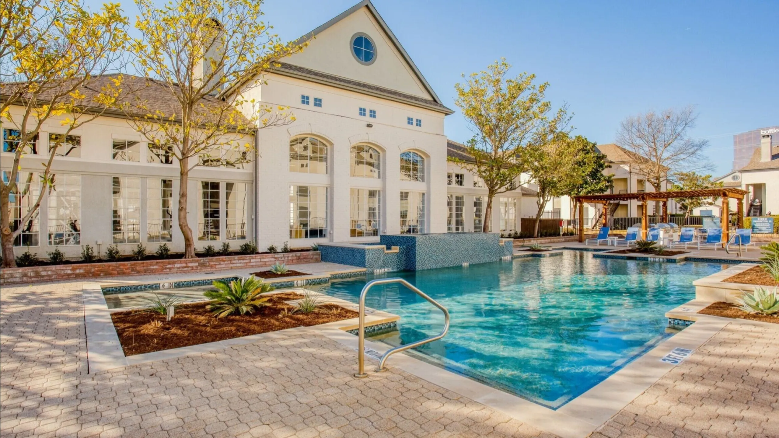 Luxurious apartment complex featuring a sparkling swimming pool, elegant white brick architecture, and a charming pergola with sun loungers, surrounded by lush landscaping.