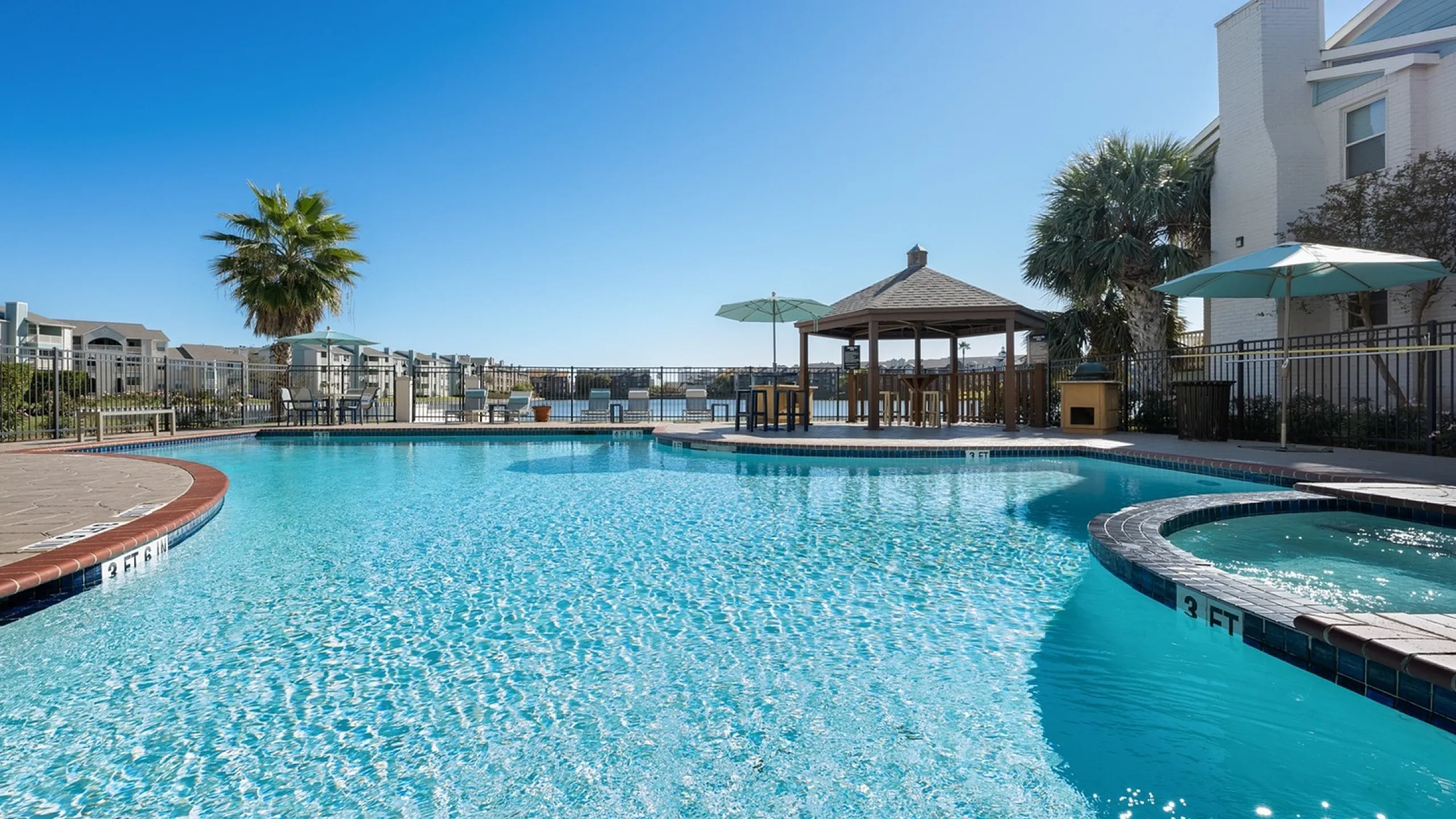 Inviting outdoor pool area with crystal-clear water, surrounded by cozy lounge chairs, palm trees, and a charming gazebo under a clear blue sky.