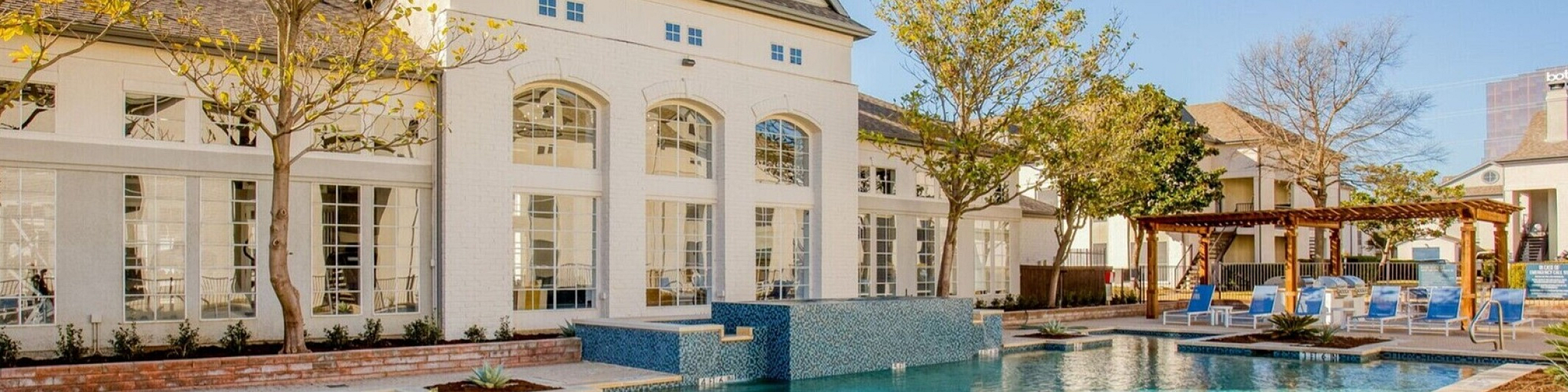 Luxurious apartment complex showcasing a sparkling swimming pool, elegant brick architecture, and lush landscaping under a clear blue sky.