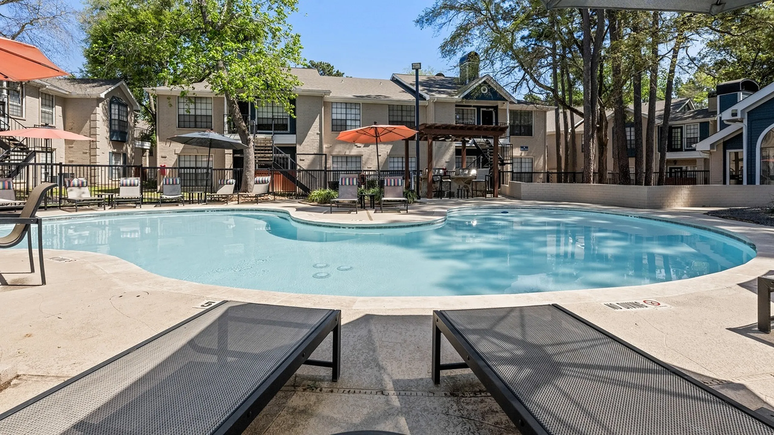 Inviting apartment complex with a sunlit pool area, surrounded by lush greenery and comfortable lounging spaces.