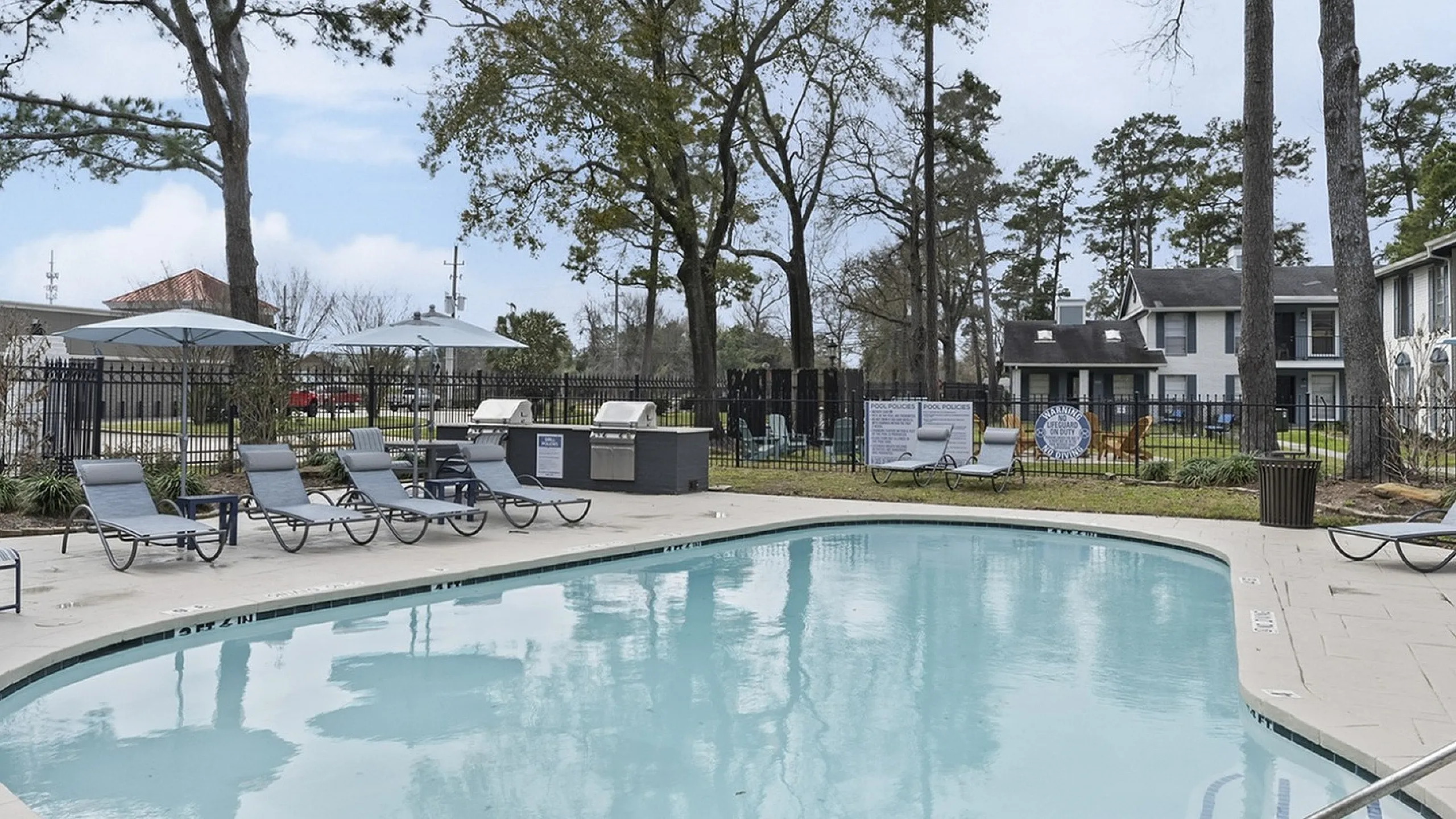 Inviting outdoor pool area with stylish seating and blue umbrellas, surrounded by lush greenery and modern apartments.