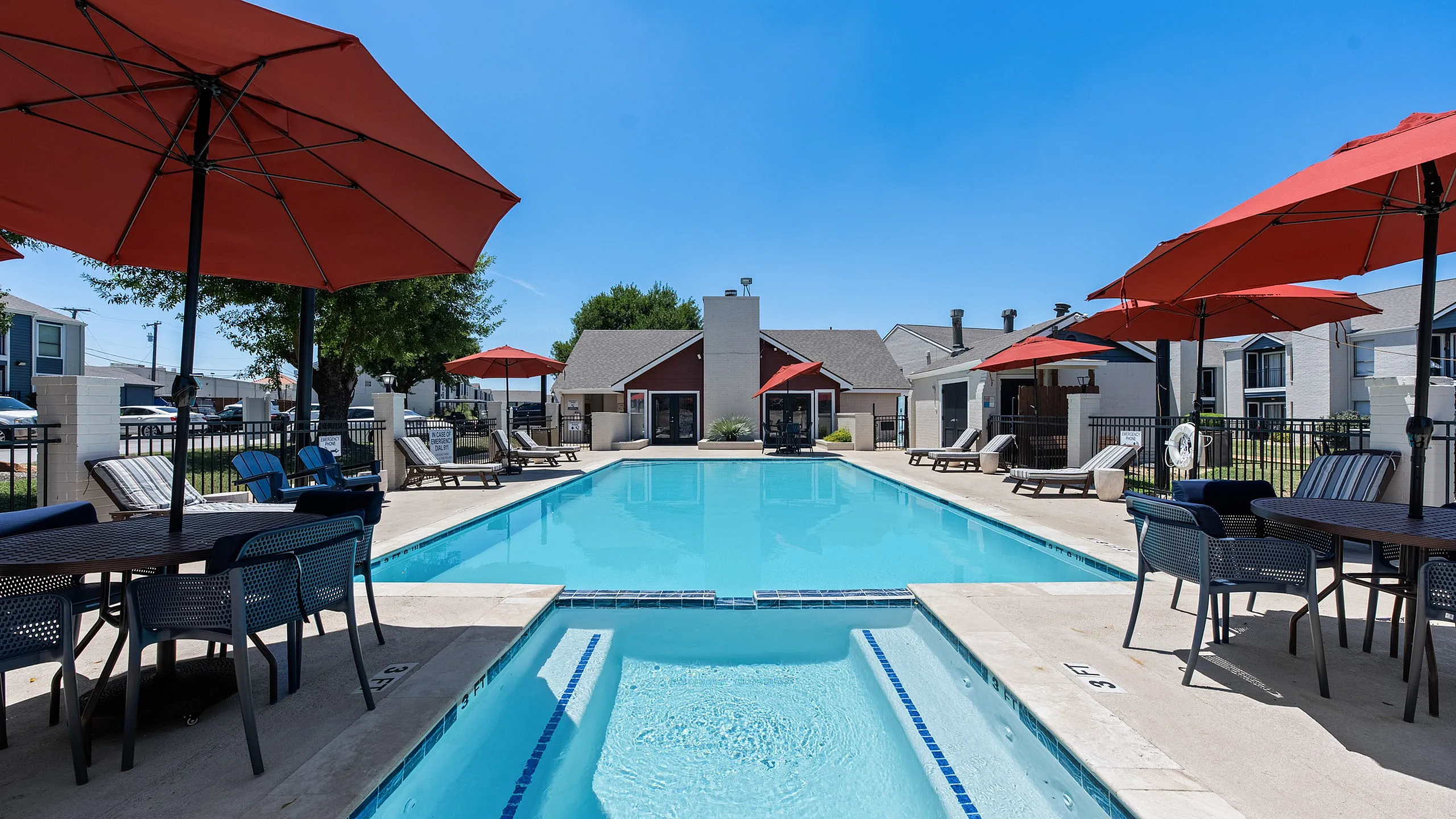 Inviting outdoor pool area with vibrant red umbrellas, surrounded by modern lounge chairs and a stylish clubhouse under a clear blue sky.