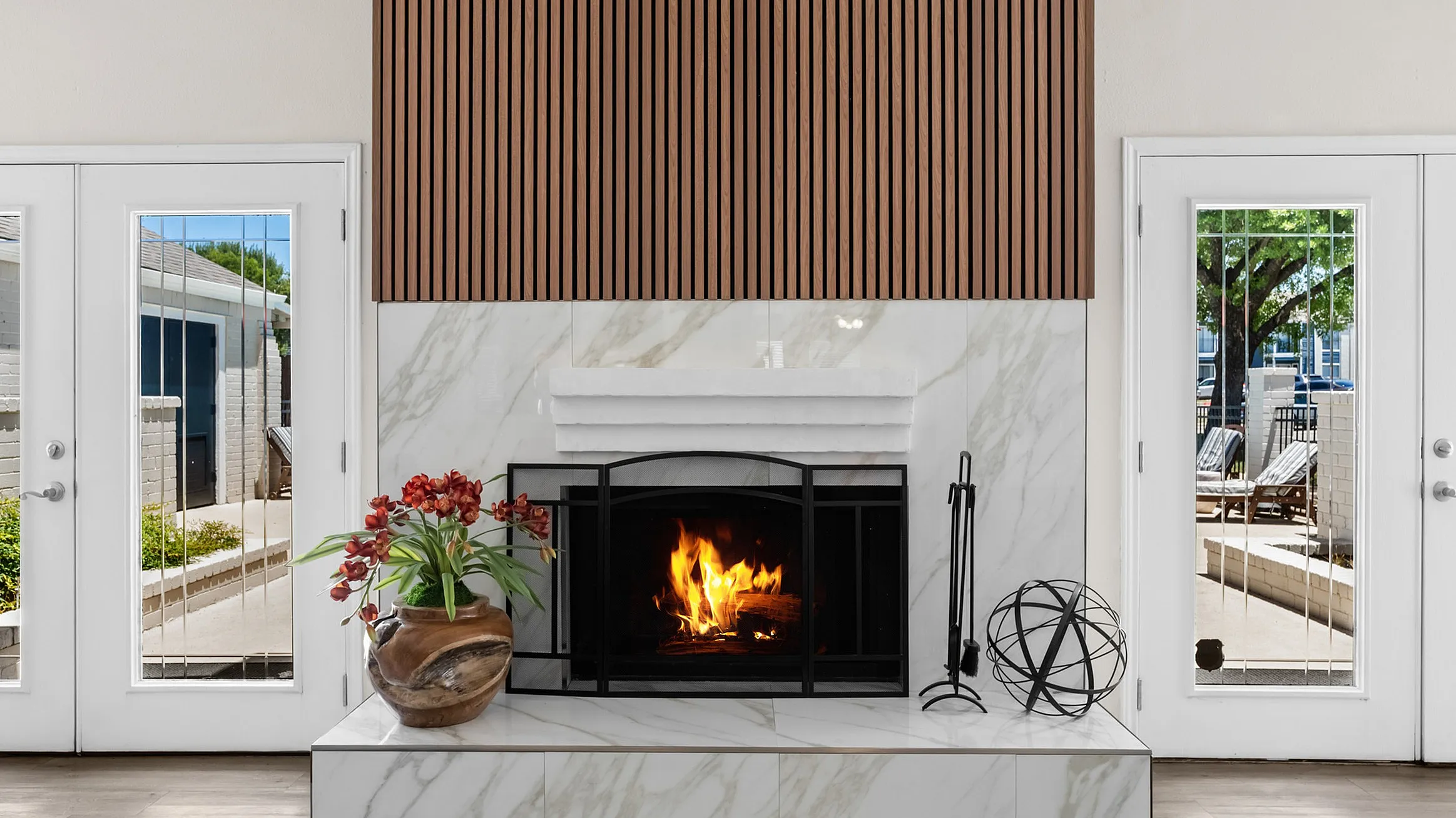 Elegant living room with a modern marble fireplace, cozy fire, and stylish wooden accents, enhanced by natural light from large glass doors.