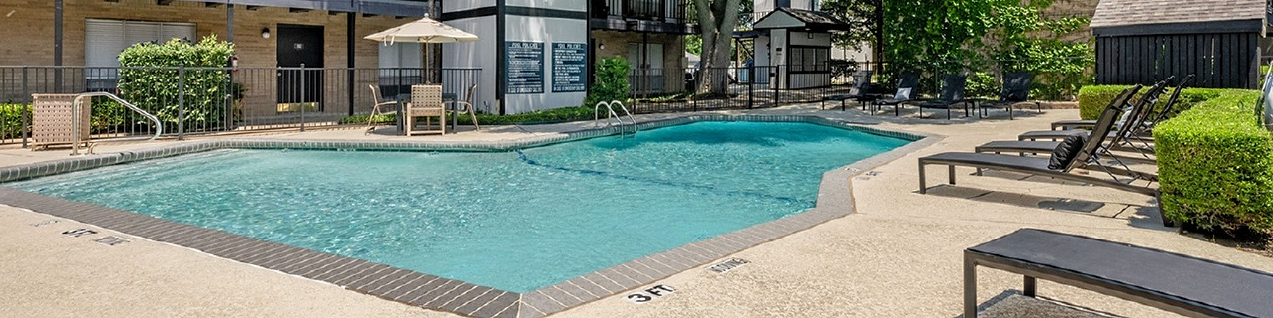 Inviting apartment complex pool area with lounge chairs, lush greenery, and serene outdoor setting under clear blue skies.