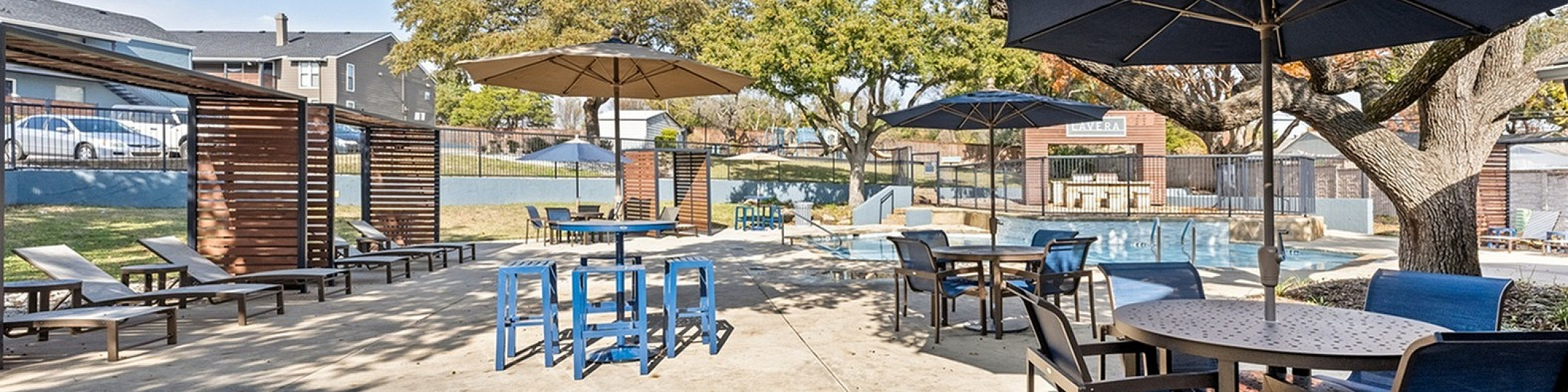 Chic outdoor patio with poolside seating, blue sun umbrellas, and shaded lounge chairs under lush trees.