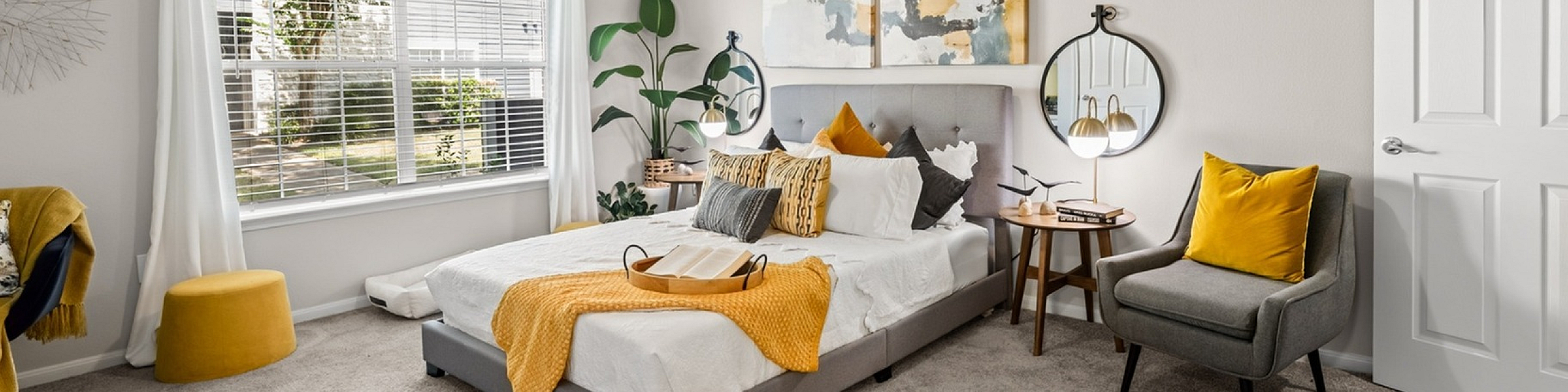 Bright and inviting bedroom featuring a modern ceiling fan, large window with lush outdoor view, stylish decor with yellow accents, and cozy seating area.