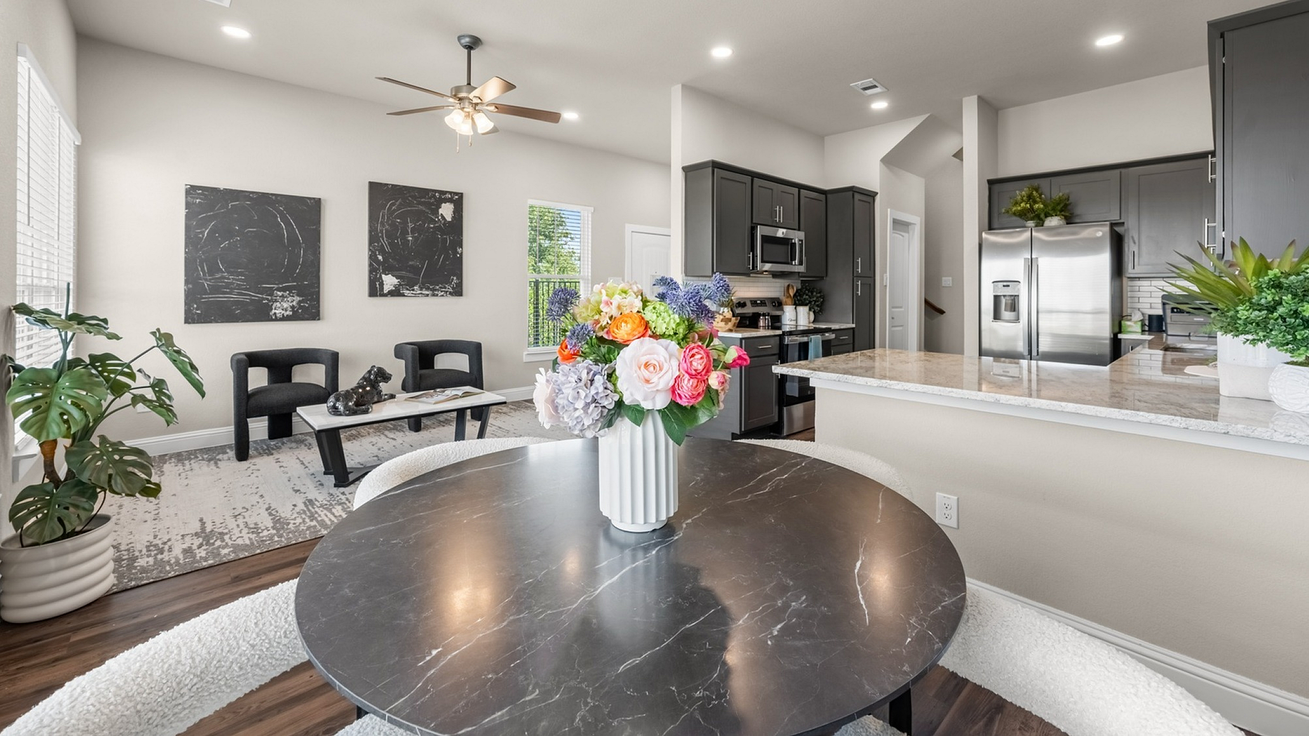 Elegant open-plan living area featuring a modern kitchen with sleek dark cabinets and stainless steel appliances, alongside a cozy dining space adorned with a vibrant floral centerpiece.